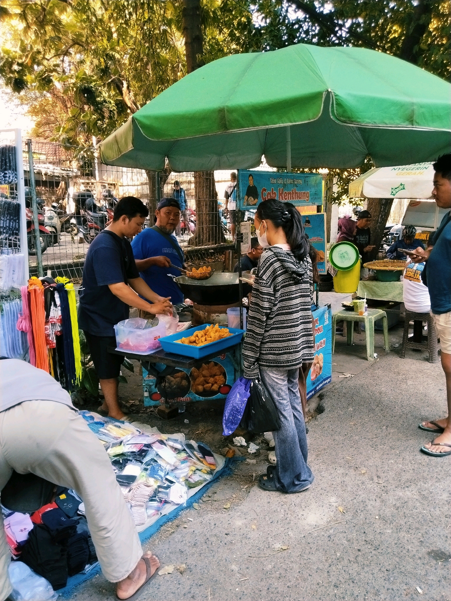 This image depicts a vibrant street scene in Surabaya, Indonesia, likely during the daytime under a clear, sunny sky. The focus is on a food stall operated by two men, with a young woman in the foreground. One man, wearing a blue t-shirt and a cap, is actively frying food in a large wok under a green umbrella. The other man, in a dark blue t-shirt, is preparing food at the stall, which displays a variety of fried snacks, including what appears to be "Tahu Walik & Balac Goreng" (turnip fritters and fried shrimp paste) and "Cob Kenthung" (a type of fried snack), as indicated by the text on the stall's signage.
In the foreground, a young woman wearing a face mask and a striped hoodie is standing, holding a blue plastic bag. To her left, a person in light-colored pants and sandals is browsing a display of clothing or accessories laid out on the ground. Behind the food stall, several motorcycles are parked, suggesting this is a busy area. Another stall with a white umbrella and a "Sprite" logo is visible in the background, indicating the presence of other vendors. The overall atmosphere suggests a bustling market or street food area, typical of urban Indonesian settings. The text visible on the stall and the "Sprite" umbrella provides specific details about the food and brands available.