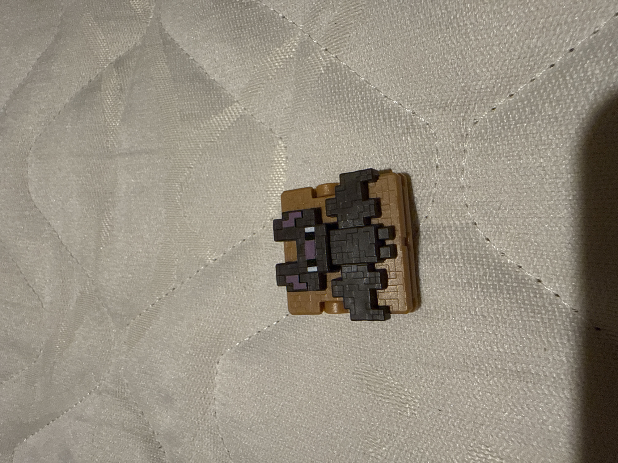 A small, pixelated toy resembling a creature from the video game Minecraft is centered in the frame. The toy depicts a dark-colored, bat-like figure, constructed from dark grey and black pixelated blocks. It features purple and white pixel details on its face or body. This figure is affixed to a flat, light brown, block-like base.
The toy rests on a light-colored, possibly off-white or light grey, quilted fabric surface, likely a mattress or bedspread. The fabric has a distinct texture, and the quilting creates raised, geometric patterns across its surface. Tiny, dark specks, possibly dust or lint, are scattered on the fabric. A soft shadow is cast to the right, suggesting indoor lighting. There is no activity taking place, and no text is visible within the image. The setting appears to be an indoor, domestic environment in Sofia, Bulgaria.