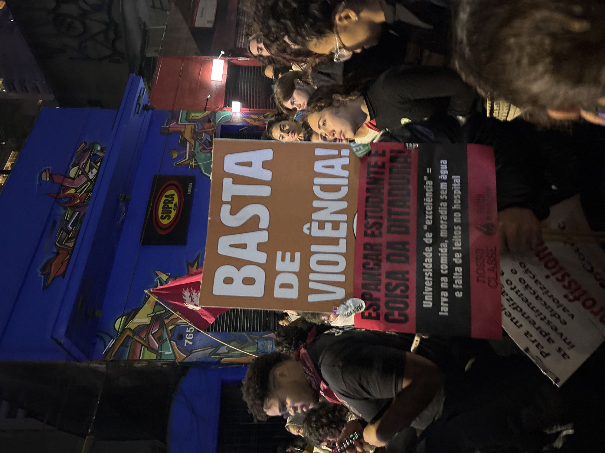 The image shows a nighttime protest in São Paulo, Brazil. A group of people are holding signs with protest messages. One large sign reads "BASTA DE VIOLÊNCIA!" (ENOUGH OF VIOLENCE!). Another sign reads "ESPANCAR ESTUDANTE É COISA DA DITADURA" (BEATING STUDENTS IS A THING OF THE DICTATORSHIP) and describes conditions at the university as "larva in the food, housing without water, and lack of beds in the hospital." The protest appears to be taking place on Augusta Street, as indicated by an "AUGUSTA" sign in the background.
A blue building with graffiti and a "SUPRA AUGUSTA" sign is visible behind the protesters. A red flag with a graphic and the number "765" on it is also visible.