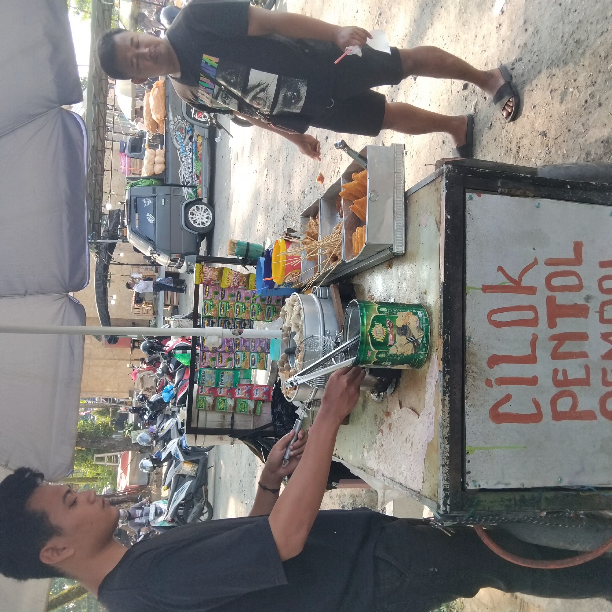 The image depicts an outdoor scene in Banjarbaru, Indonesia, likely a street food stall. A young man in a black t-shirt and dark pants is standing behind a food cart, holding tongs over a metal pot filled with food items. The cart has various food items displayed in metal trays, including what appear to be fried snacks on skewers and triangular-shaped food items. There are also packaged snacks and beverages displayed on the side of the cart. In the background, several motorcycles are parked, suggesting a busy area. A grey van with colorful graphics is parked nearby, and other individuals are visible in the background, some standing and some on motorcycles. A second man, wearing a black t-shirt with a graphic print and black shorts, stands to the right of the food cart, holding a white cup with a straw. The ground is dirt and gravel. The overall impression is of a bustling street market or a busy roadside food vendor. The signage on the cart appears to be in Indonesian, with the words "CILOK PENTOL" clearly visible, indicating the type of food being sold.