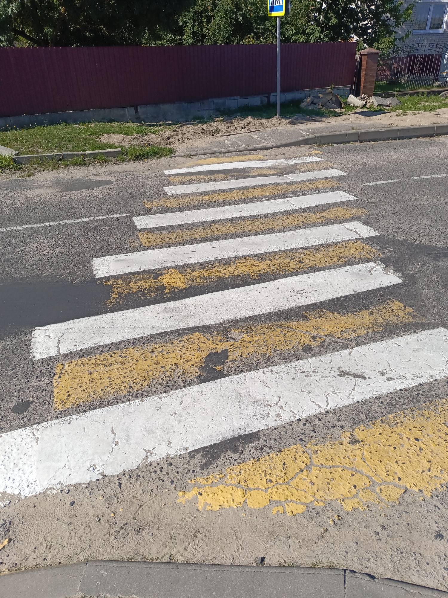 The image shows a pedestrian crossing on a road in Ivatsevichy, Belarus. The crossing has alternating white and yellow stripes, with some of the paint showing signs of wear and tear. The road surface is asphalt. To the left of the road, there is a dirt verge with some sparse grass and a concrete curb. Further back, there is a dark red metal fence and some trees. To the right of the road, a pedestrian crossing sign with a blue background and white pictograms is visible, along with a brick pillar and a wrought iron fence. The scene appears to be a typical urban street environment. There are no people visible in the image.