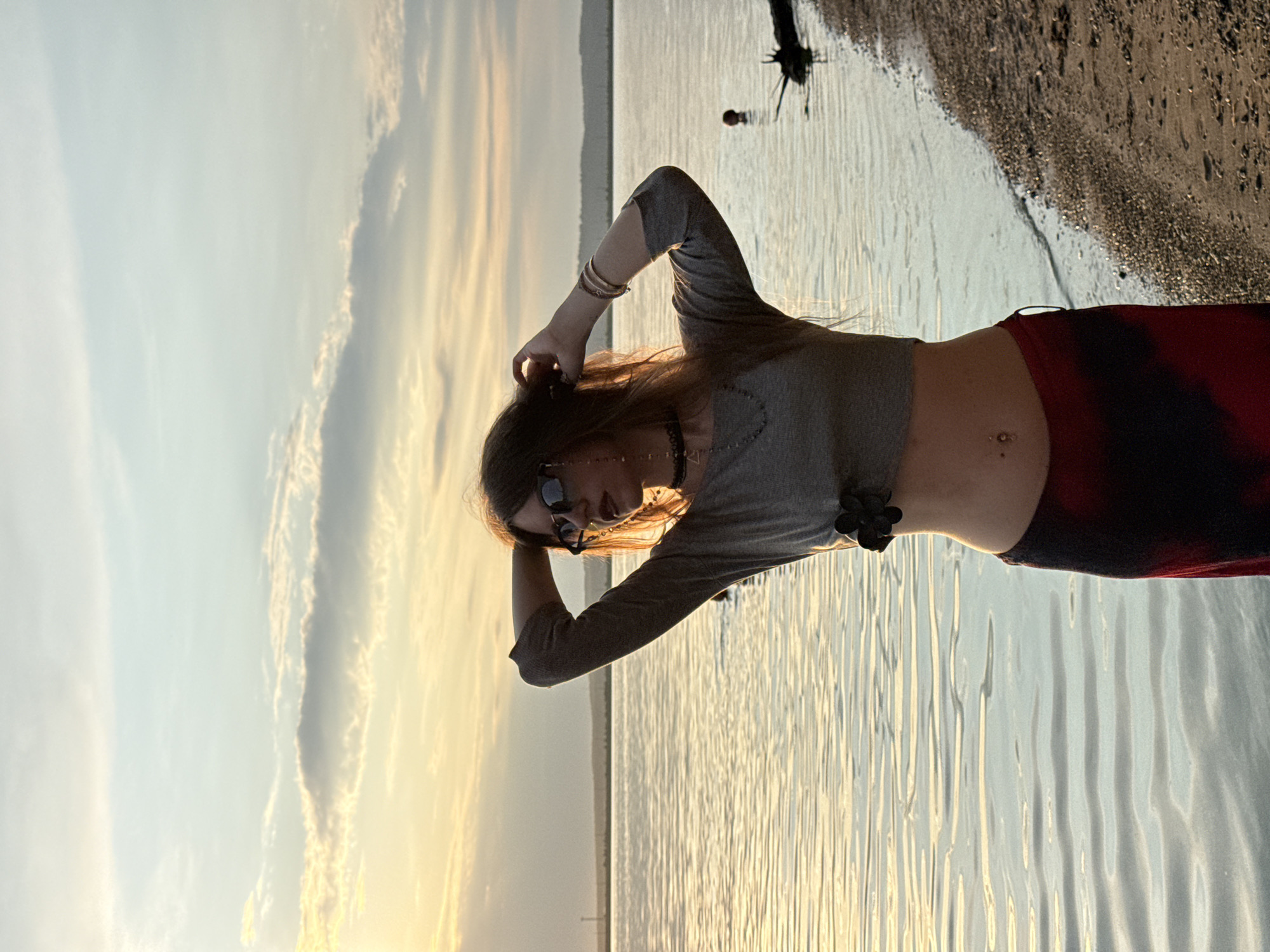 A young woman with long brown hair stands on a sandy shore by a body of water. The sun is setting, casting a warm glow on the scene. She is wearing a grey crop top and a red and black patterned skirt. She has on sunglasses and is holding her hands up to her hair. The water is calm, with gentle ripples. The sky is filled with soft clouds, tinged with orange and yellow from the setting sun. In the distance, a treeline is visible on the horizon. The image captures a serene moment of a woman enjoying the natural beauty of her surroundings.