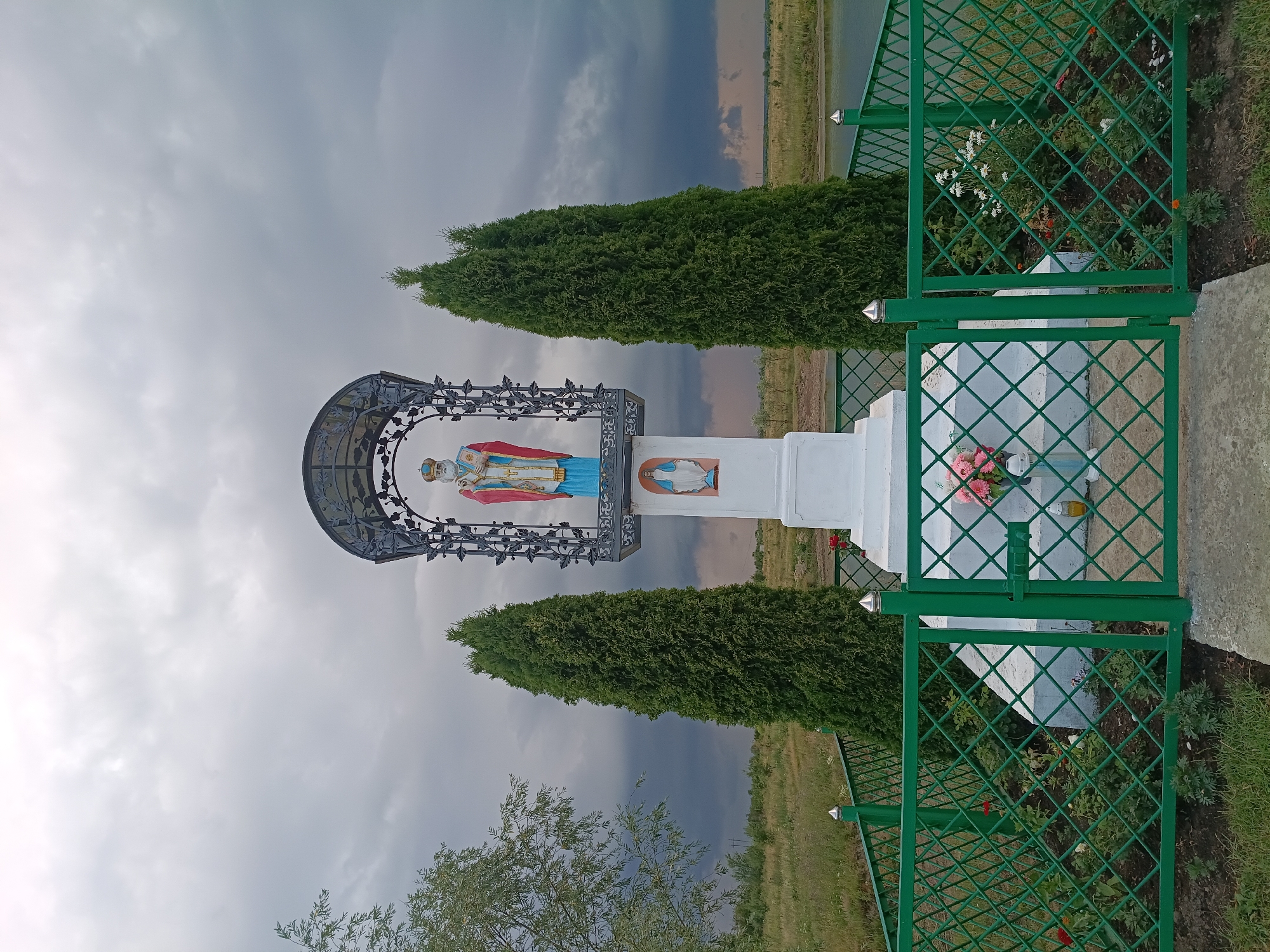 A statue of Saint Nicholas stands atop a white pedestal within a metal enclosure. The statue is adorned with a blue robe, a red cloak, and a crown. Below Saint Nicholas, an image of the Virgin Mary is depicted. The pedestal is surrounded by a green metal fence with a diamond pattern. Two tall evergreen trees flank the statue. The scene is set outdoors with a body of water and a field in the background, under a cloudy sky. This monument is located in the city of Litynia, Ukraine.