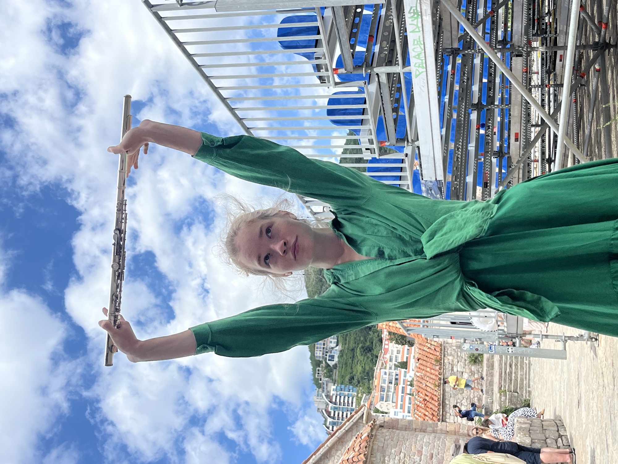 A young woman with blonde hair tied back stands outdoors, holding a silver flute above her head with both hands, looking upwards. She wears a long-sleeved, green, draped dress cinched at the waist. Behind her, a blue sky with white clouds is visible. To her right, a metal structure with blue stadium seating is partially visible, suggesting an outdoor venue or seating area. In the background, buildings with terracotta roofs and modern structures are nestled on a green hillside, characteristic of Budva, Montenegro. The foreground shows a cobblestone pathway and a stone wall, with other people in the distance. The overall scene appears to be a sunny day in a historic coastal city.
