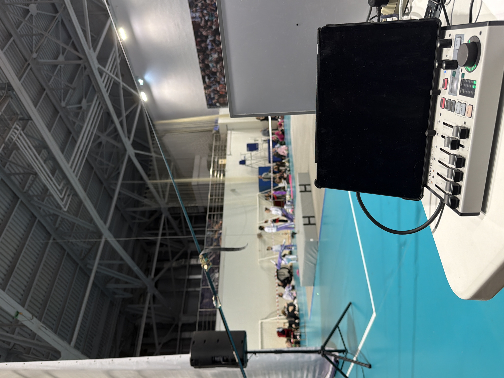 This image depicts an indoor sports gymnasium in Perm, Russia, captured from a control station setup in the foreground.
The foreground features a white table holding a grey and white "FLOW 8 Digital Mixer" with multiple faders, buttons, and a large rotary knob, indicating an audio or production control point. An electronic tablet, with a black screen and its case, is positioned upright above the mixer, likely serving as a control interface or monitor. Behind this setup, a rectangular white screen or board partially obscures the background.
To the left of the control station, a black speaker is mounted on a tripod stand, facing towards the center of the hall. A thin blue cable stretches horizontally across the mid-ground, originating near the speaker and extending towards the right.
The mid-ground reveals a light-teal colored sports court with white demarcation lines. A raised, grey, padded platform, bearing two distinct black "H" letters on its side, serves as a performance or activity area. Several young individuals, predominantly female, are present around and on this platform. One individual, dressed in a light purple uniform, appears to be in the midst of a gymnastic or performance pose on the platform, with others observing or waiting nearby. Additional individuals are seated on the floor or on low benches behind the performance area. White, net-like structures and miscellaneous equipment are visible in this zone.
The background shows the extensive interior of the gymnasium. The high ceiling features exposed metal trusses, corrugated panels, and a network of utility pipes and conduits. Multiple bright lights illuminate the space. The far wall displays a large photo mural depicting a dense crowd, possibly spectators at a sporting event. An elevated level or balcony with a railing is visible along the far wall, suggesting an upper viewing area. Various white support columns and wall sections complete the structural elements of the large hall.