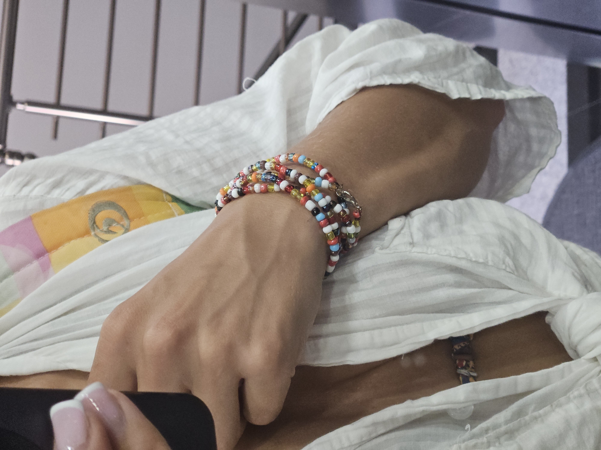 A close-up, medium shot captures a person's right arm and chest area, draped in a loose, white, crinkled fabric garment. The arm is adorned with multiple colorful beaded bracelets, primarily featuring shades of red, white, blue, orange, and yellow, with a silver clasp visible on the outermost bracelet. A portion of the person's left hand, with manicured nails, is seen holding a dark object, likely a phone, on the left side of the frame. The background is blurred, suggesting an indoor setting with a metallic railing and grey walls. The person's skin tone is medium-toned.