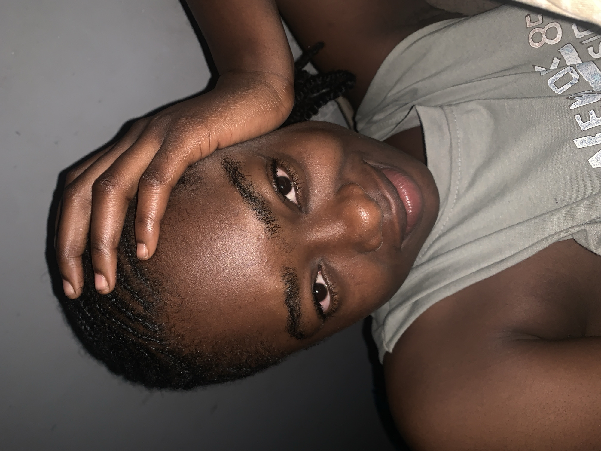 An upward-facing, slightly rotated close-up portrait shows a person with dark skin and short, neat braided hair, possibly cornrows. The subject is lying down, looking directly at the camera with clear, open eyes and a slight, subtle smile. One hand, with visible fingernails, rests gently on the side of their head, creating a relaxed pose.
The person is wearing a light grey or faded green t-shirt. Visible white text on the shirt includes numbers like "80" and partial words such as "NEW Y" (likely part of "NEW YORK") and "WE A". There is subtle facial hair or stubble visible on the cheeks and jawline. The lighting is dim, suggesting an indoor setting, with highlights catching on the skin, giving it a healthy sheen. The background is a plain, dark, solid color, possibly a wall or surface, indicating a simple indoor environment. There are no clear outdoor or weather cues. The overall mood appears calm and contemplative. This scene takes place in Rafin Par, Nigeria.