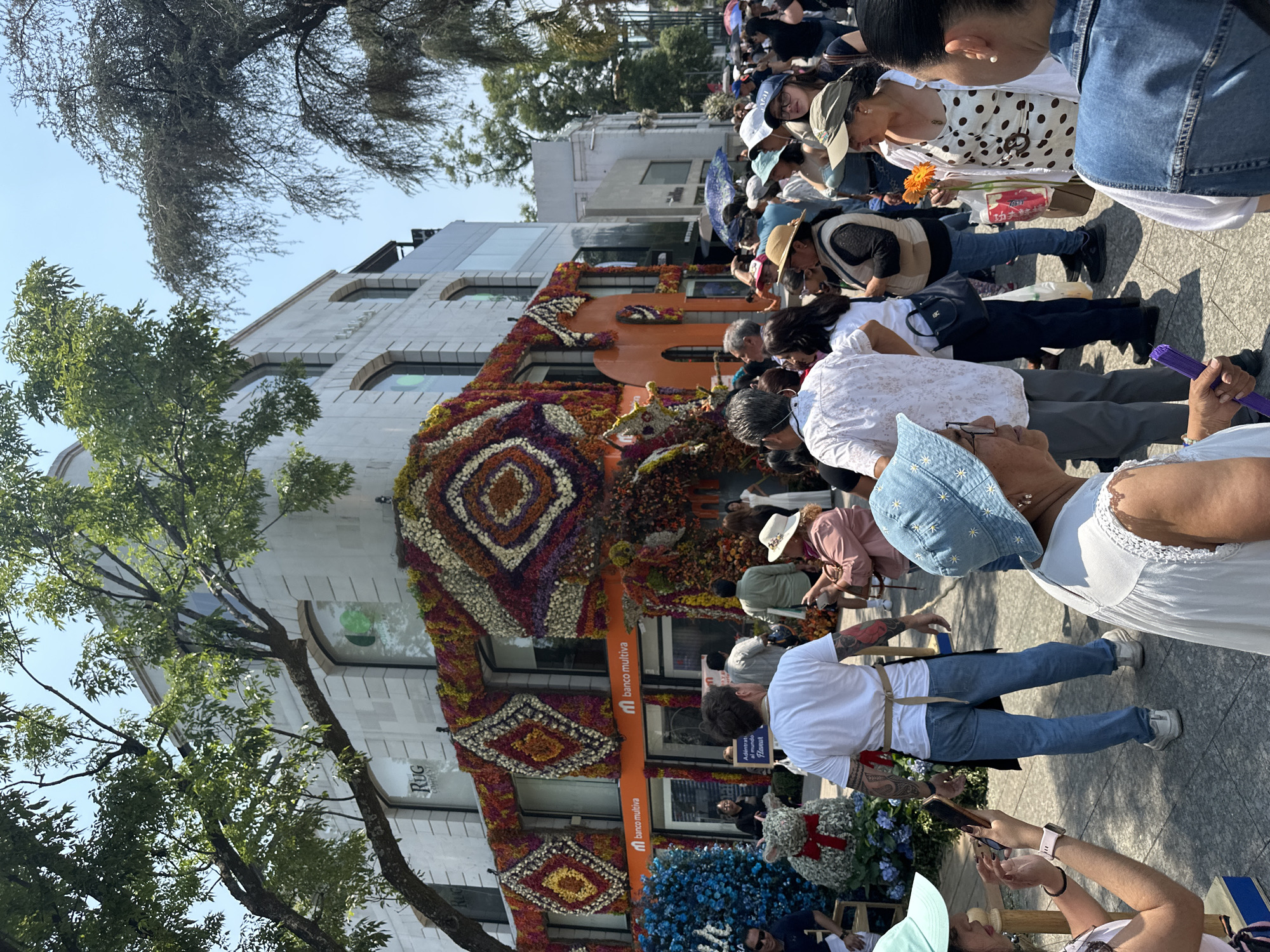 The image shows a building facade in Mexico City, Mexico, elaborately decorated with floral arrangements in a geometric pattern, likely for a festival or event. The building appears to be a branch of "banco multiva." Crowds of people are viewing and photographing the display. Trees partially obscure the building. The sky is blue with some clouds. The street is paved and appears to be in a busy urban area. People are dressed in casual clothing. Some individuals are holding flowers.