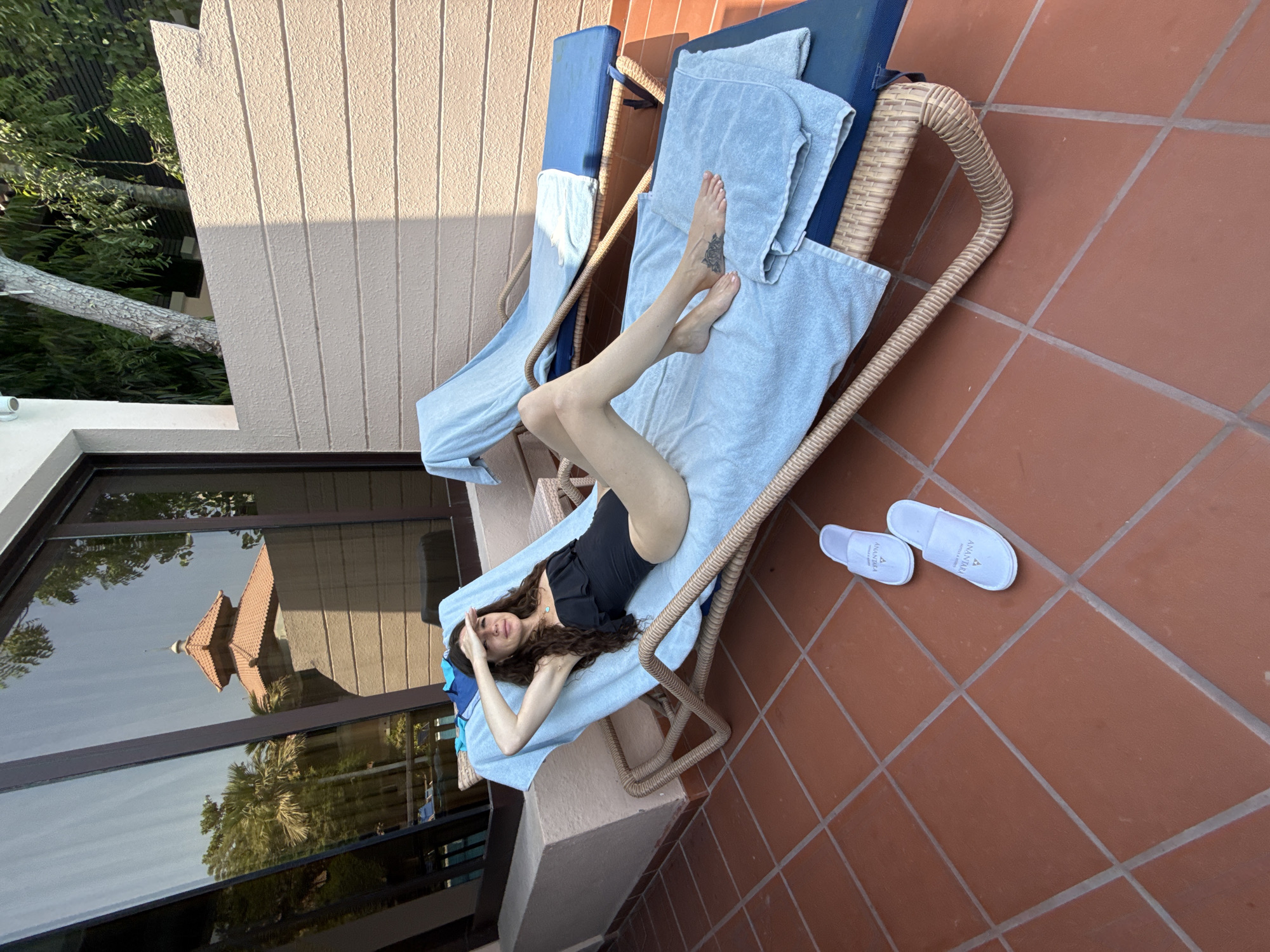 A woman with long, wavy brown hair is sunbathing on a chaise lounge on a tiled patio. She is wearing a black one-piece swimsuit and is holding her hand up to block the sun from her eyes. She is lying on a light blue towel. Another chaise lounge, also with a blue towel, is visible behind her. Two white slippers are on the tiled floor in front of the chaise lounge. The background shows a building with large windows reflecting trees and a structure with a red-tiled roof. The scene appears to be outdoors, possibly in a resort or hotel setting.