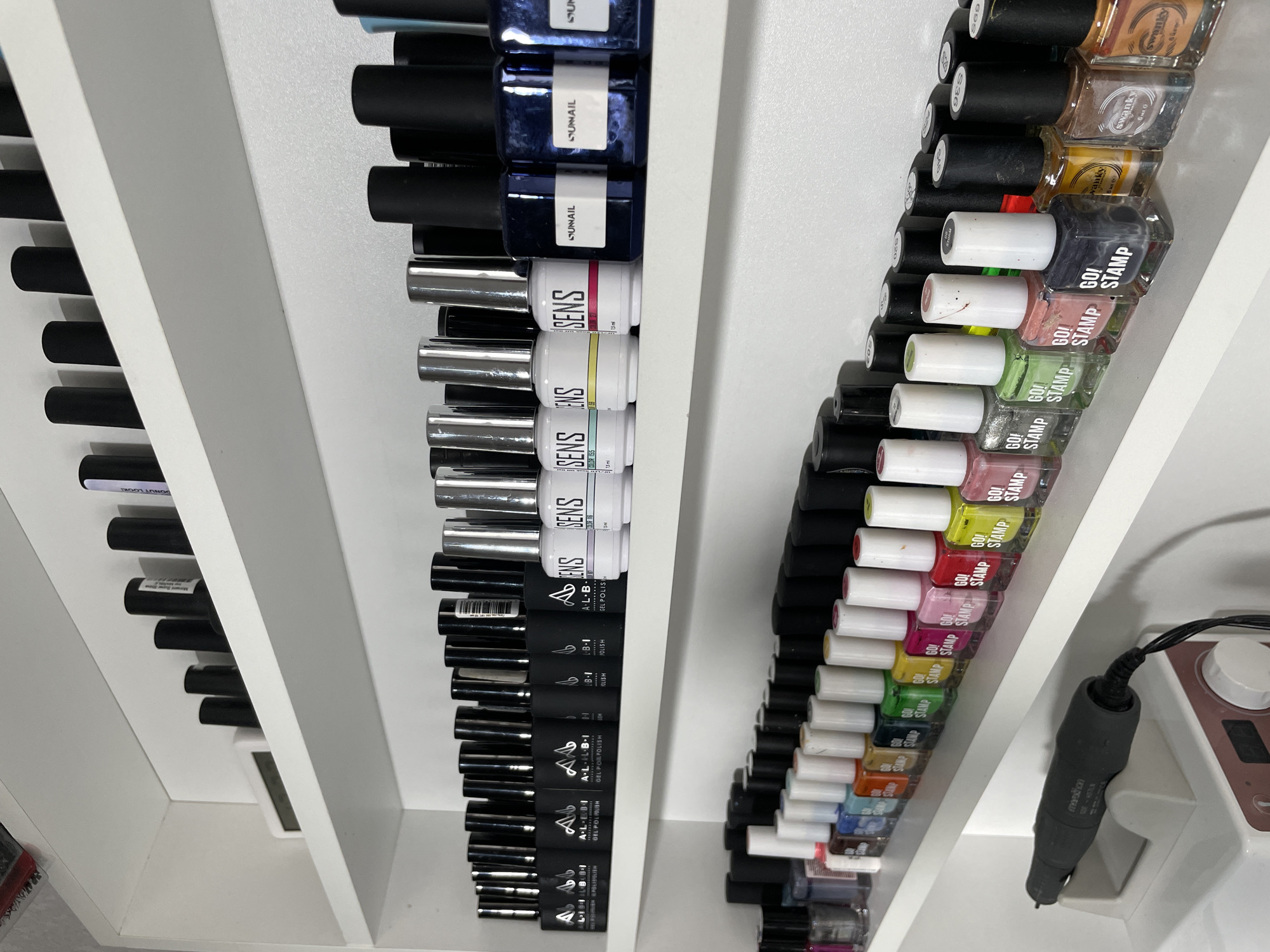 This image shows three rows of nail polish bottles stored on white shelves, likely inside a beauty salon or nail technician's workspace. The brands visible include "A.L.B.I GEL POLISH," "SENS," "SUNAIL," "GO! STAMP," and "swanky." The bottles are arranged vertically, with a variety of colors visible. A nail drill is visible in the right side of the picture. It is impossible to determine the exact location in Izhevsk, Russia.