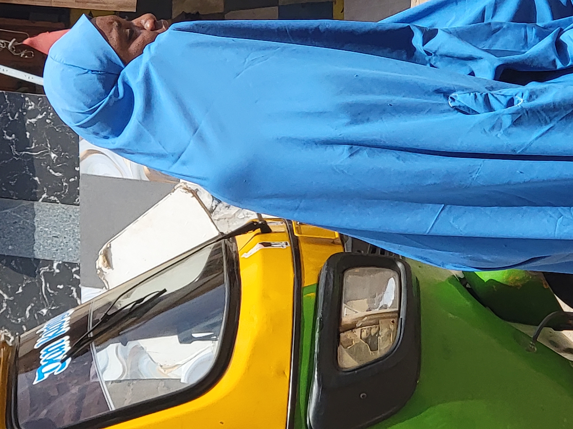 An elderly Black male, wearing a blue head covering and draped in a blue sheet, lies horizontally on a yellow and green auto rickshaw (tuk-tuk) in Maiburiji, Nigeria. The vehicle is parked near a gray tiled surface and a white object. The scene is sunlit.