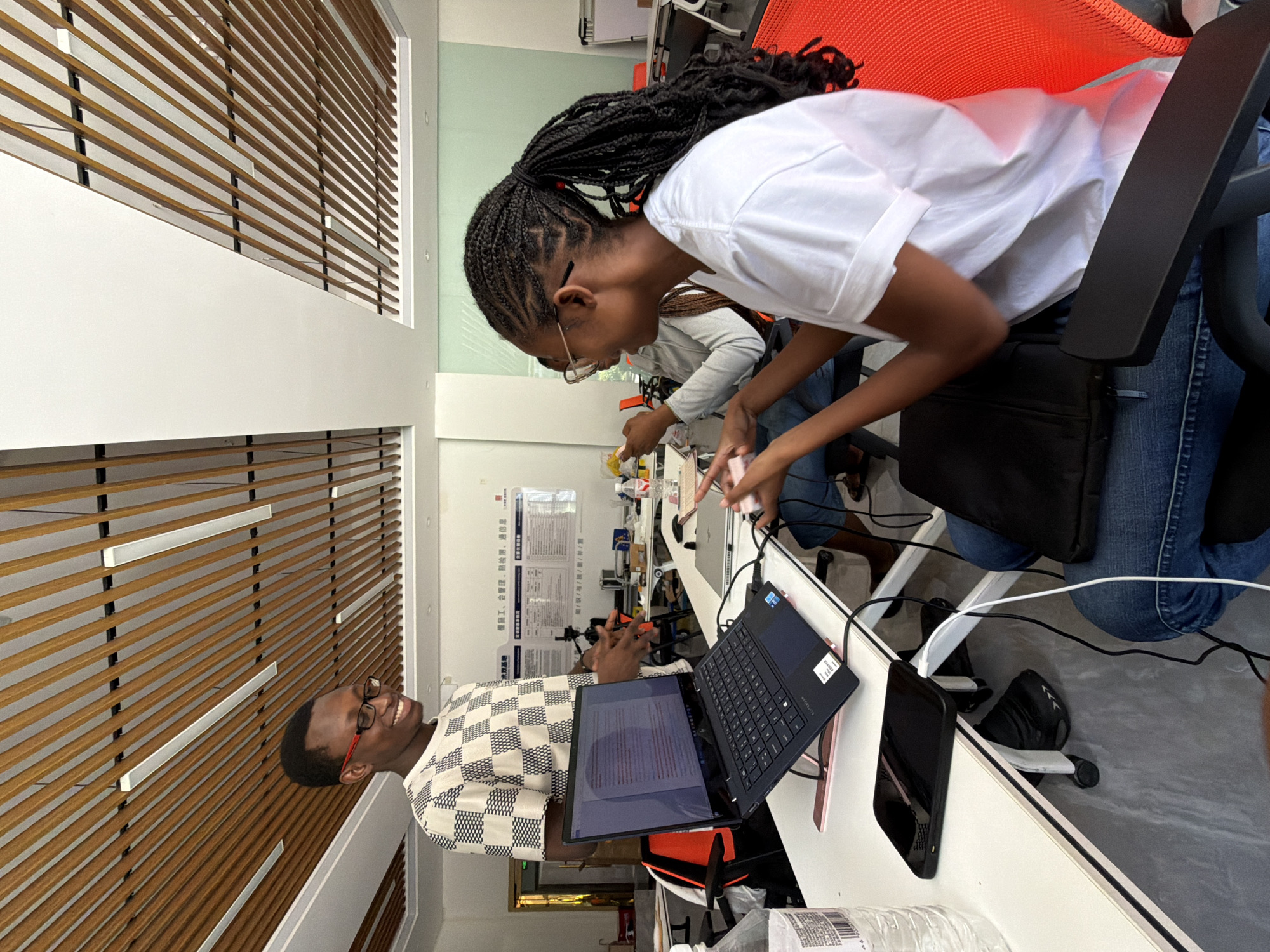 The image shows an indoor scene, possibly an office or co-working space, with a man and two women. The man, wearing glasses and a checkered shirt, stands to the left, gesturing towards a laptop on a table. The laptop is open and displays text. To the right, a woman with braided hair, wearing a white t-shirt and jeans, is seated and looking at a phone or small device. A second woman, partially visible, is seated behind her, also engaged with a device. The environment features a modern ceiling with wooden slats and recessed lighting. A whiteboard with text is visible on the wall in the background, and desks with chairs are arranged in the space. A bottle of water is on the table in the foreground. The overall impression is one of a collaborative or work-related setting.