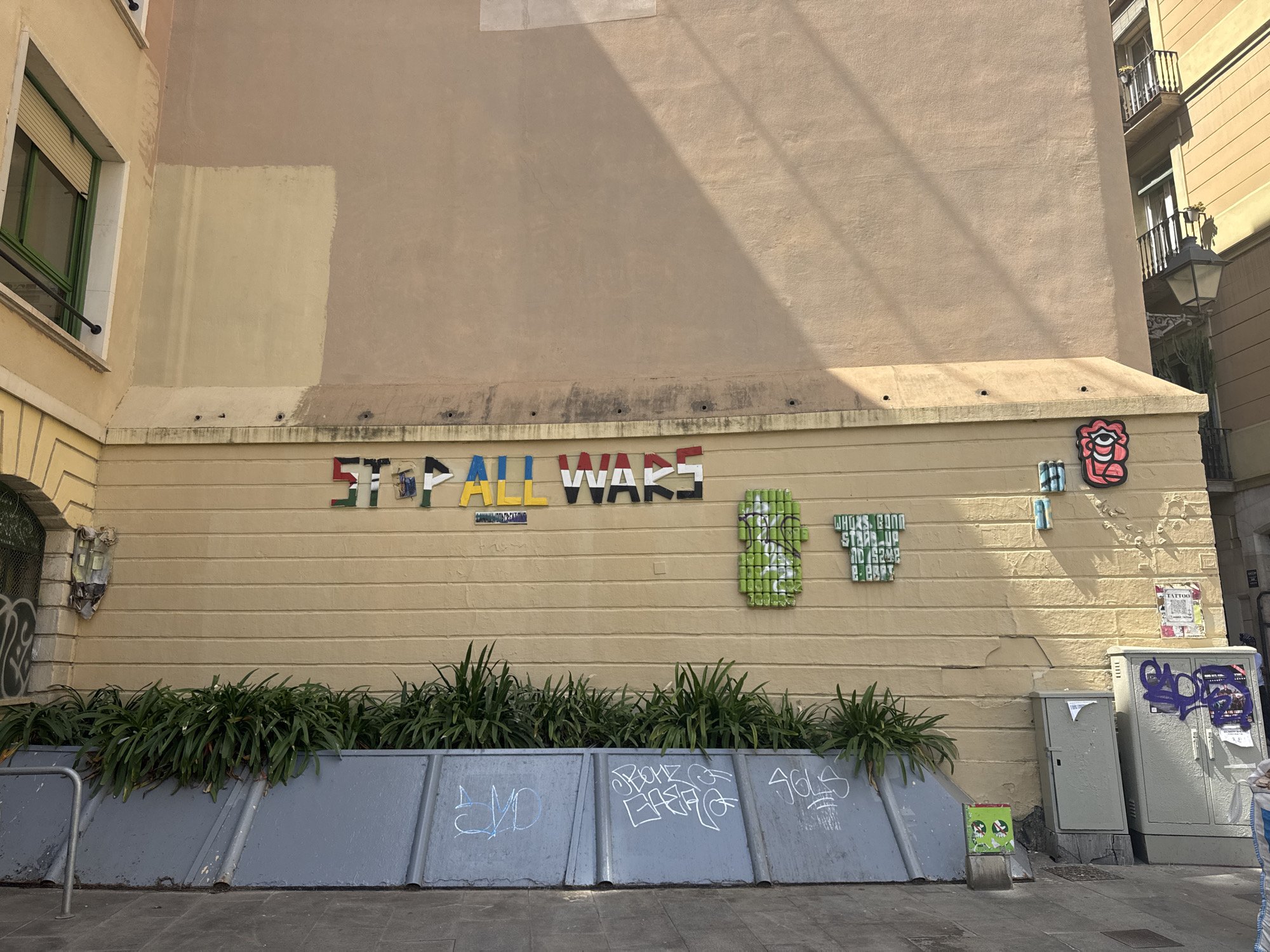 This media file captures a vibrant urban street scene in Barcelona, Spain, centered on a light beige building wall adorned with multiple pieces of street art under bright daylight.
The main subject is a building wall, which serves as a canvas for various artistic expressions. Prominently displayed is a large, colorful, metallic-looking text piece spelling "STOP ALL WARS". To its right, another artwork made from what appears to be green crushed cans forms the phrase "WHO'S GONNA STAND UP AND SAY ENOUGH", accompanied by a smaller "NO" in blue and white letters. Further right, a stylized red and white figure resembling an eye or mouth is affixed to the wall.
Below the wall, a long planter filled with lush green foliage runs horizontally. In front of the planter are grey corrugated metal barriers, heavily marked with various graffiti tags, including one that appears to say "SVD". The pavement or sidewalk is visible in the foreground. To the far right, two grey utility boxes stand, also covered in graffiti (one with purple tagging) and partially torn posters, one of which has "TATTOO" visible. A small green box with a cartoon face rests on the ground beside the barriers. On the left side of the image, part of another building is visible, featuring a window with a green frame and closed blinds. In the background on the right, other buildings with balconies and a black street light are visible.
The time of day is clearly sunny, with distinct shadows cast across the upper part of the wall from structures off-frame, indicating bright sunlight and clear weather. There are no people visible in the scene. The street art conveys a strong anti-war message, adding an emotional and political layer to the urban landscape.