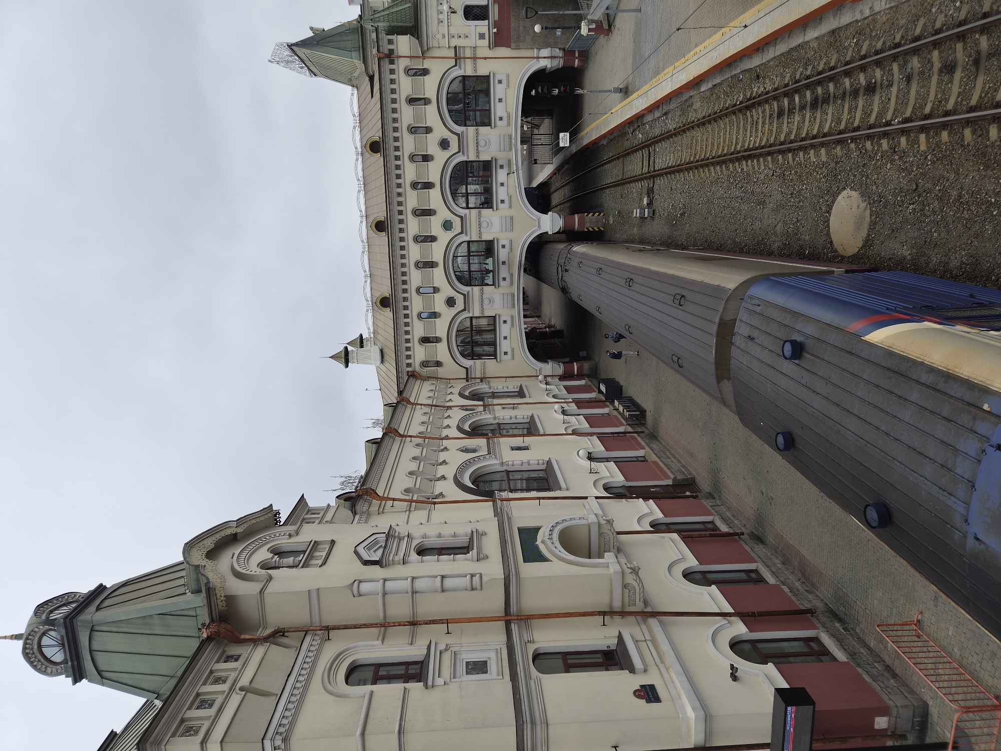 This is a high-angle, eye-level shot of the Vladivostok railway station in Russia. The station building, with its distinctive cream and maroon facade and ornate architectural details, dominates the left side of the frame. A green-roofed tower with a domed top is visible. A skybridge, also cream-colored with arched windows, connects the two parts of the station over the train tracks. A train, with a long gray and blue carriage, is parked on the tracks. Two people are visible on the platform to the left of the train, and another two are on the skybridge. The weather appears overcast. The train tracks run diagonally through the scene, with a yellow-lined platform visible on the right.
