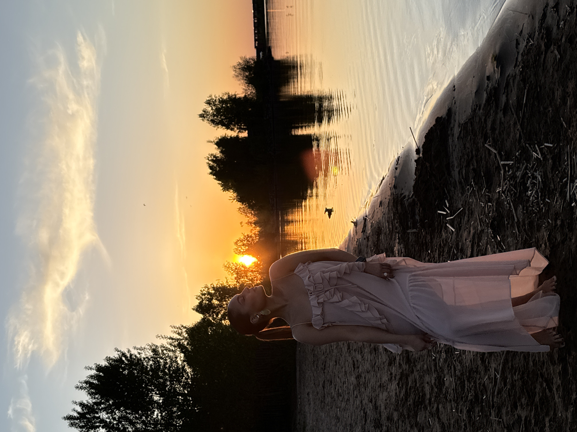 A woman in a long, light-colored dress is standing on a sandy beach at sunset. The scene is illuminated by the orange and golden light of the setting sun, which is visible in the background and reflected in the water. Trees line the horizon. The location is likely the city of Chaikovsky, Russia, based on the prompt. The environment consists of a sandy beach, calm water, and a sunset sky. The woman is standing still, possibly posing for a photo.