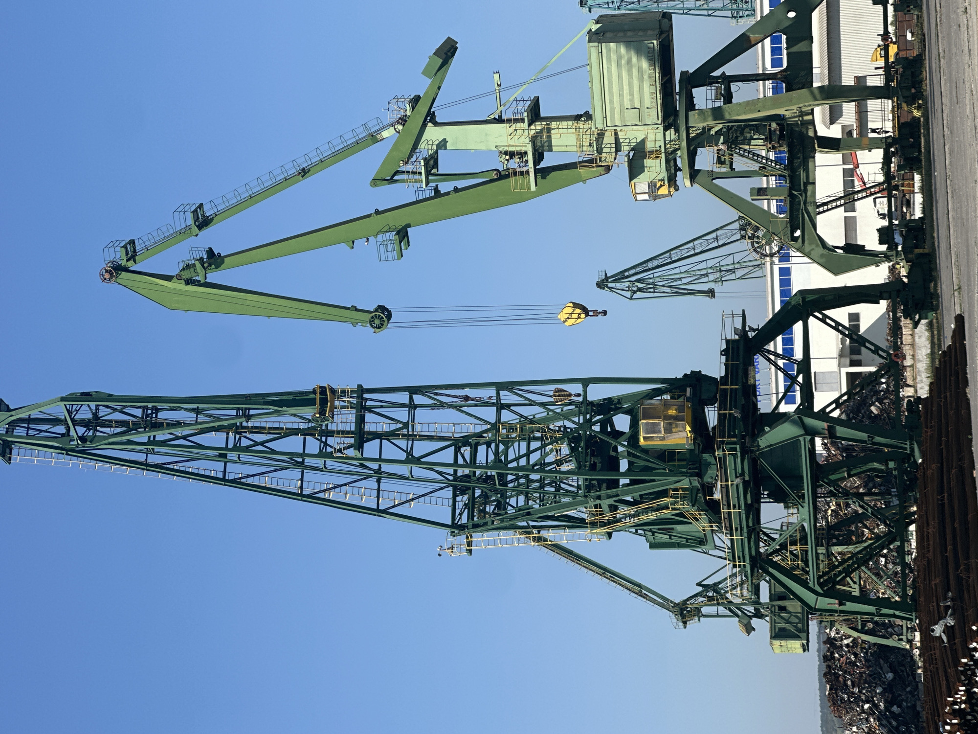 The image shows an industrial port scene with multiple large green cranes against a clear blue sky. In the foreground, a pile of rebar is visible. Behind the rebar, a large pile of scrap metal is situated. Two prominent cranes dominate the center of the image, with their tall, latticed structures reaching high into the sky. One crane, on the left, is taller and more slender, while the crane on the right is wider and has a more robust construction. Both cranes have operators' cabins. A yellow hook is suspended from one of the cranes. In the background, there are several industrial buildings and what appear to be more cranes of varying sizes. A yellow truck is also visible in the middle ground. The overall impression is of a working port or scrapyard.