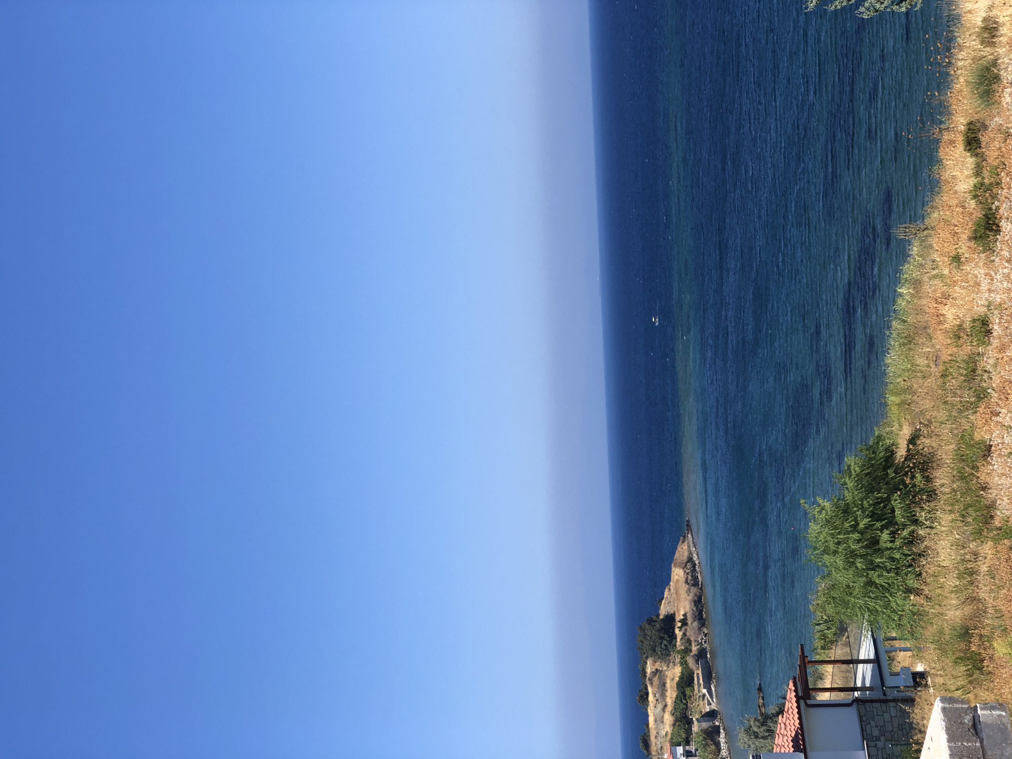 This is an eye-level, medium shot photograph of a coastal scene in Karaburun, Turkey. The upper portion of the image is dominated by a clear, bright blue sky. Below the sky, the deep blue sea stretches to the horizon, with a faint haze visible where the sky meets the water. A small white boat is visible in the distance on the sea.
The foreground of the image features dry, yellow grass and sparse vegetation. On the left side, a white building with a red tiled roof and a stone foundation is partially visible, featuring a covered porch area with wooden pillars. Behind the building and to the right, a small, dense green tree stands out against the dry landscape.
The middle ground of the image shows a rocky, dry coastline with scattered green trees and shrubs. The sea recedes into a bay with calmer, lighter blue waters closer to the shore. The overall impression is a sunny, tranquil, and arid coastal environment.