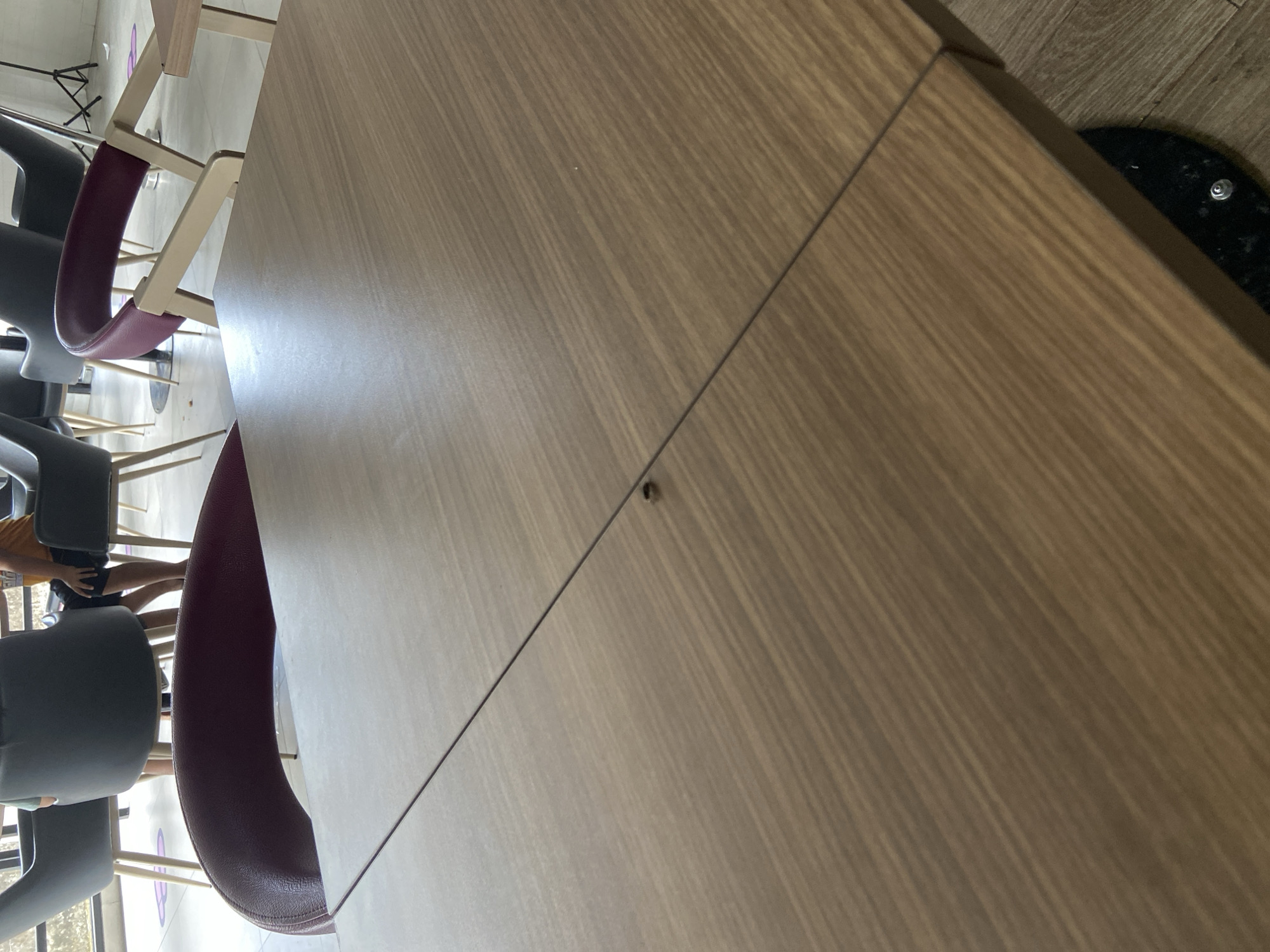 The image shows a close-up of a wooden table in a cafe. The table has a visible seam running down the middle. A small, dark object, possibly a fly or a piece of debris, is located on the surface of the table near the seam. In the background, several chairs are visible, some are dark grey, and one is burgundy. The floor appears to be tiled. The setting is likely an indoor public space. There are no specific visual cues in the image that indicate the location is in Bago, Indonesia.