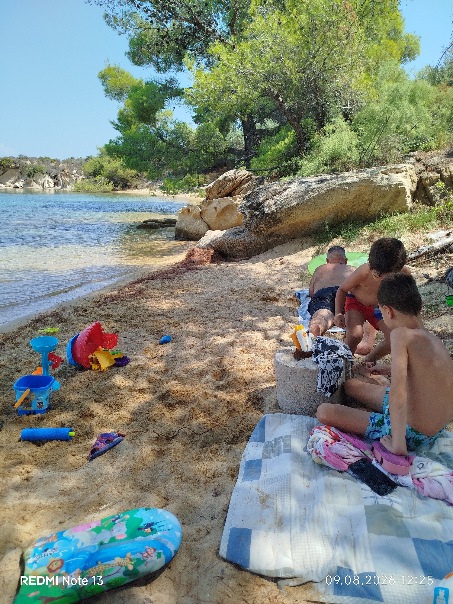 In Sithonia Municipal Unit, Greece, on a sunny day, a beach scene unfolds with children playing in the sand near the clear blue water. Three boys, aged approximately 5-10 years old, are present. One boy in red swim trunks is facing away from the camera, while another boy in blue patterned swim trunks is seated, looking down. A third boy, further back, is also facing away. They are situated on a patterned blanket next to a concrete block serving as a makeshift table. Scattered around are various children's beach toys, including buckets, shovels, and a blue water gun. In the background, a tranquil body of water meets a sandy beach lined with rocks and lush green trees, suggesting a secluded coastal area. The timestamp at the bottom of the image indicates August 9, 2026, at 12:25.