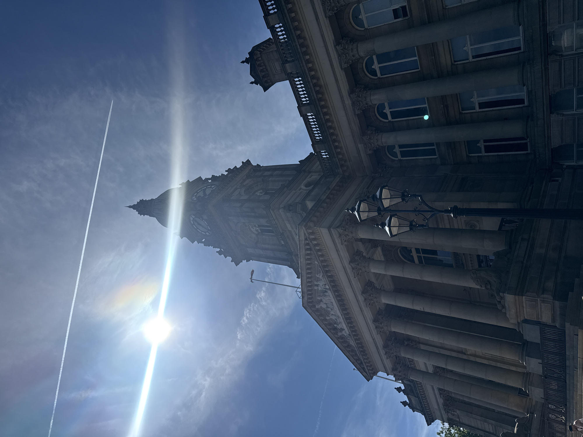 The image captures a low-angle, wide shot of the Bolton Town Hall in Bolton, United Kingdom, under a bright, sunny sky with a few wispy clouds. A prominent contrail from an aircraft stretches across the upper portion of the sky. The sun, positioned in the upper left quadrant, is intensely bright, causing lens flare and a visible rainbow-like effect.
The grand facade of the Town Hall dominates the lower and right portions of the frame. It features a classical architectural style with numerous Corinthian columns, a pediment adorned with sculptures, and rows of arched windows. A large clock tower rises above the main building, its upper section partially obscured by the sun's glare.
In the foreground, a vintage-style lamppost with three globes stands to the right of center. The stone steps leading up to the building are visible at the bottom left, with a wrought-iron fence partially framing the lower area. Stone lion sculptures are positioned at the base of the building, flanking the entrance. The overall impression is one of stately grandeur and a clear, bright day in a public square.