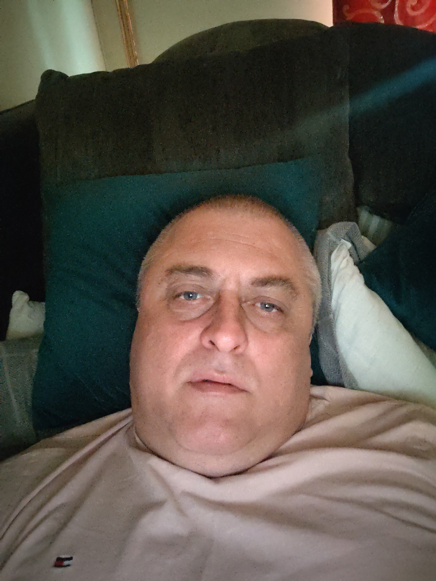 A man is lying down on a sofa with green and dark grey cushions, and a white pillow. He is wearing a light pink t-shirt with a small red and black logo on the left side of his chest. The man has short grey hair and blue eyes, and is looking towards the camera. The image is taken from a close-up perspective, showing his face and upper body. The background is dark, with some light reflecting off a surface on the right side of the image, possibly a window or a lamp. No specific location in Huntingdonshire, United Kingdom can be identified from the image.