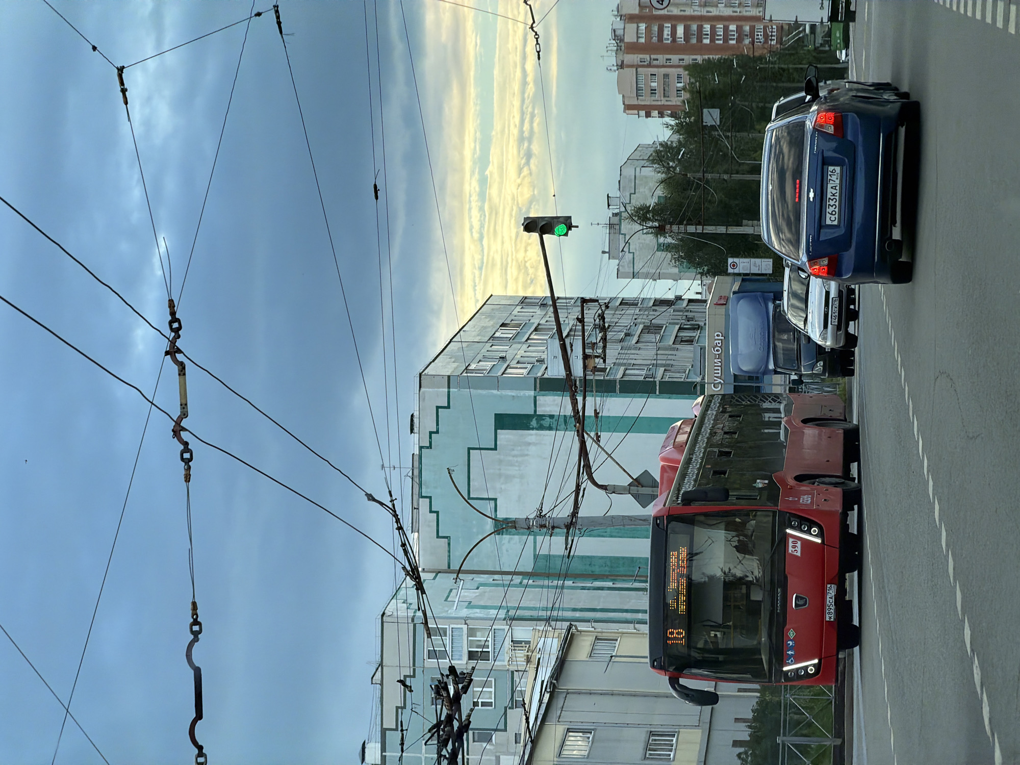 The image depicts a street scene in Kazan, Russia, with a red bus, a blue Chevrolet sedan, a blue truck, and a white car visible. The bus, numbered 18, is on the left side of the frame, facing forward. The Chevrolet sedan is in the right lane, also facing forward. A blue truck is in the left lane, ahead of the white car. A traffic light is green. The background consists of apartment buildings and overhead wires. The sky is cloudy.