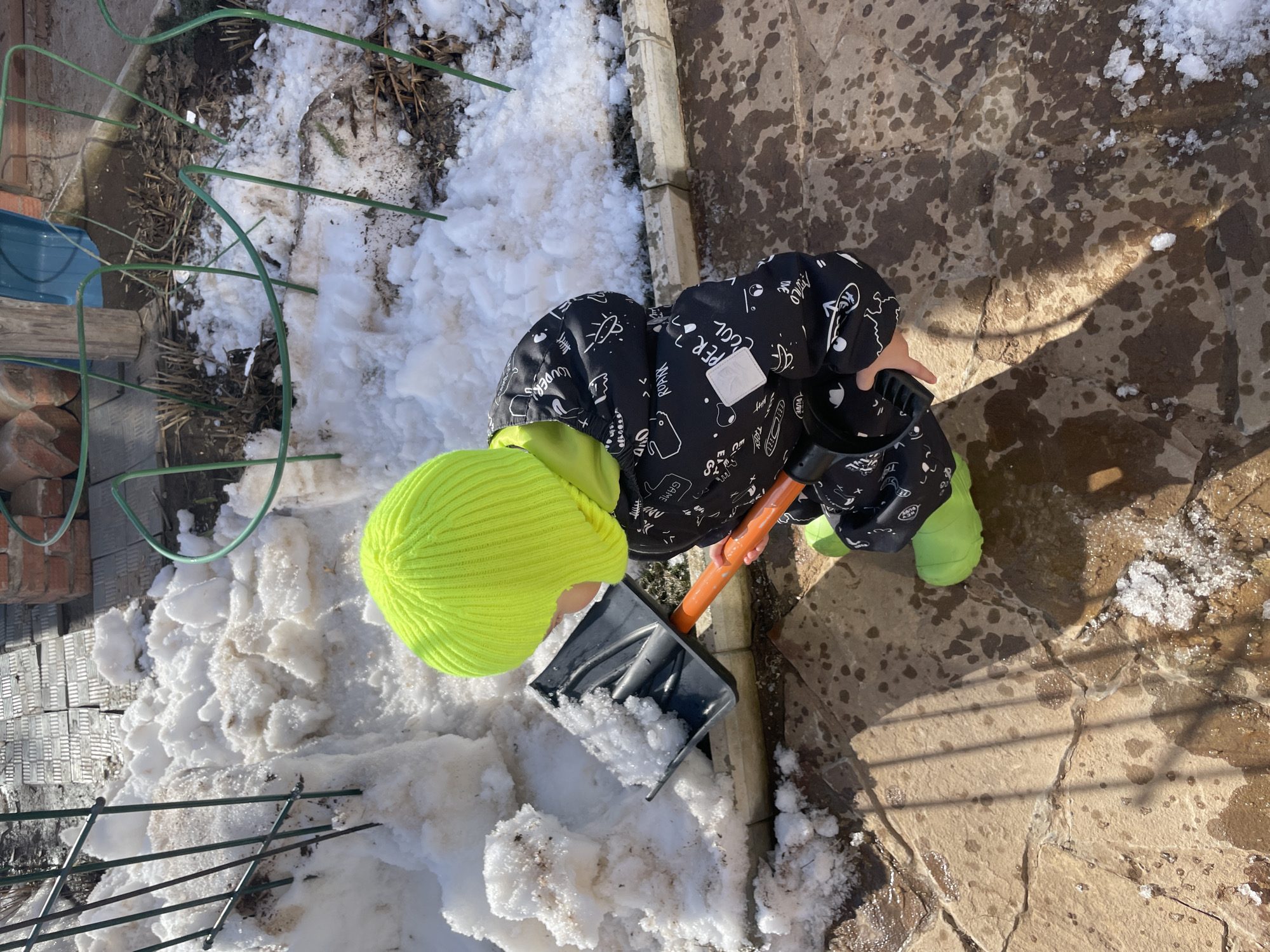 An outdoor scene in Zav'yalovo, Russia, depicts a young individual from a high-angle perspective, engaged in shoveling snow. The individual wears a bright neon yellow knitted beanie, a black snowsuit adorned with white cartoon-style graphics (including text like "GAME" and "SUPER COOL"), and bright neon green rubber boots. They hold a black plastic shovel with an orange shaft, actively scooping dirty, melting snow. The ground consists of irregular, tan-colored flagstones, extensively wet with visible puddles and patches of ice and melting snow. Strong sunlight casts distinct shadows, including linear shadows from a metal fence on the ground. To the left, a pile of partially melted, soiled snow is visible next to green metal garden fencing. To the right, another section of green metal fencing borders an area of bare, dark soil with dormant plant remnants and additional melting snow. A brick structure and a wooden post are visible in the background.