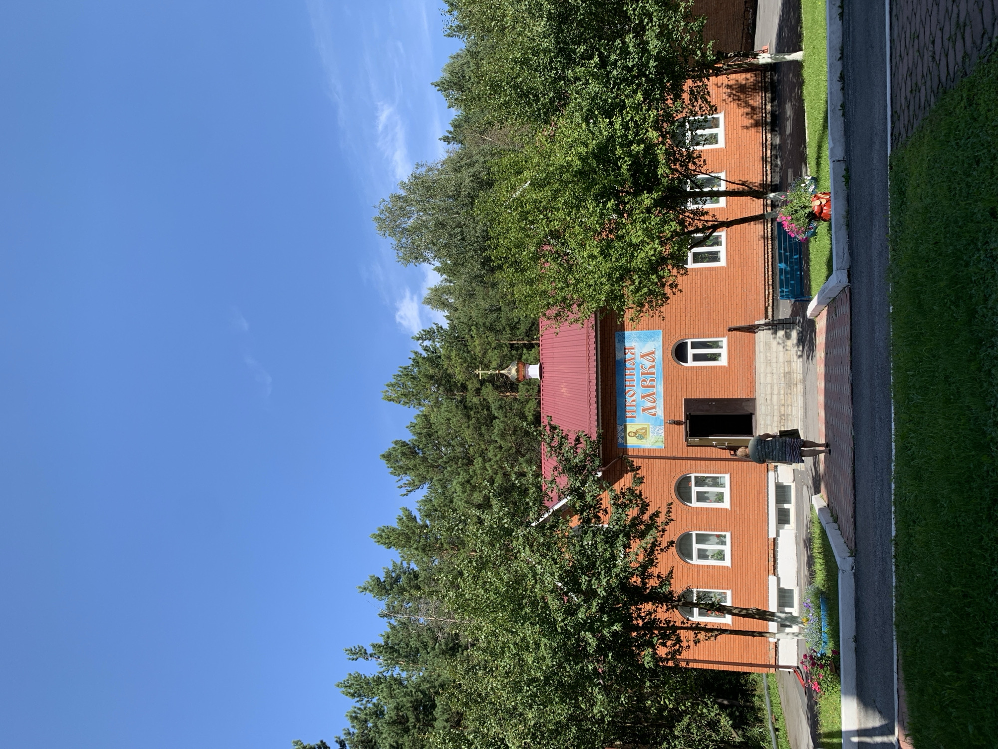 The image shows a small, red brick building with a red roof and a golden dome topped with a cross. The building appears to be a church or a chapel, possibly a part of a larger religious complex. Above the entrance, a blue sign with Cyrillic lettering is visible. The building is surrounded by lush green trees, and in the foreground, there is a paved pathway leading to the entrance. A woman in a striped dress is walking towards the entrance, carrying a bag. The sky is a clear blue with a few wispy clouds. The scene is bathed in bright sunlight, suggesting a warm day. Based on the architectural style and the context of a church building, this image likely depicts a place of worship in Prokopyevsk, Russia, possibly within a monastery or a church compound.