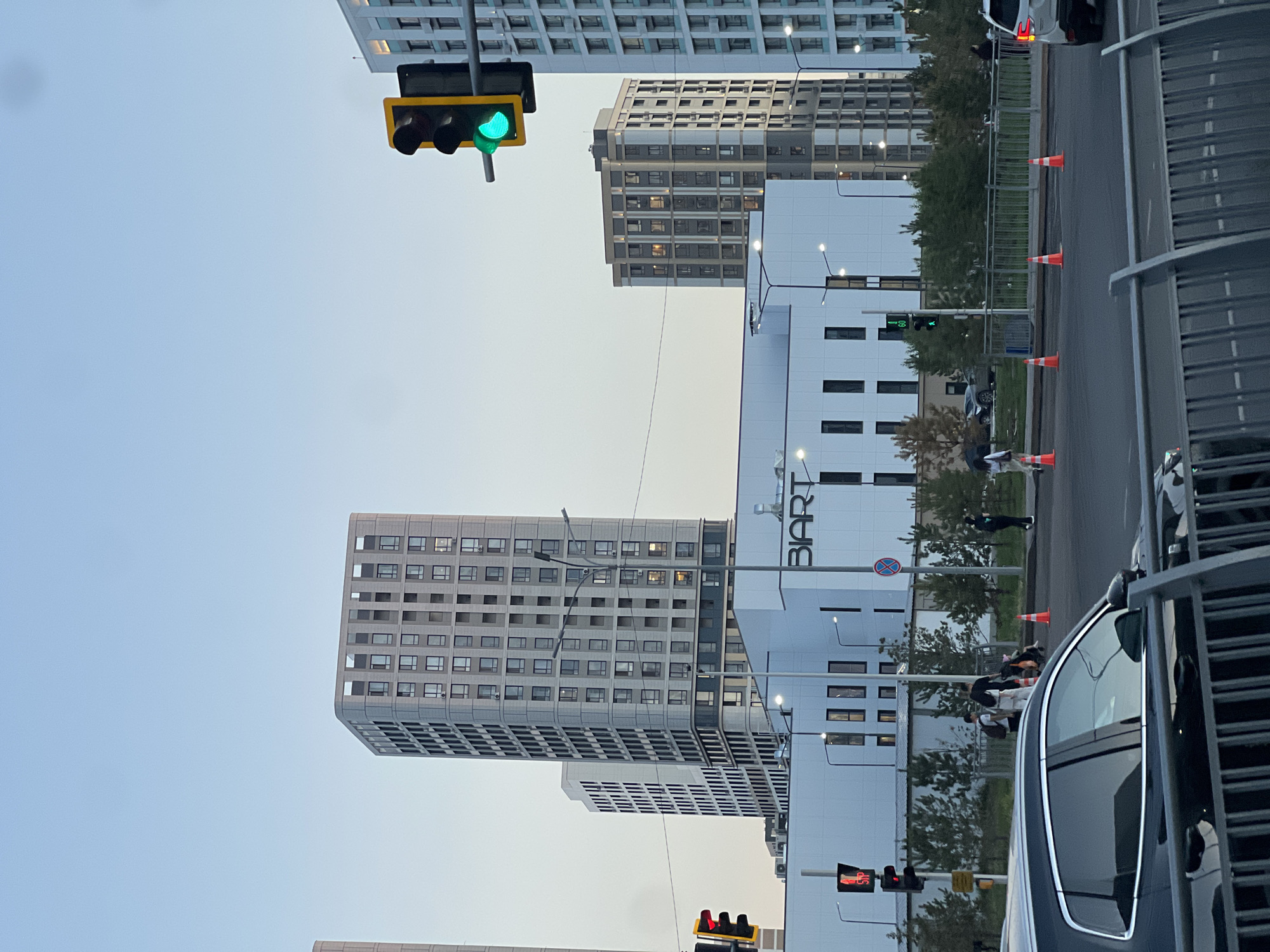 The image depicts a street scene in Astana, Kazakhstan, with a clear sky. In the foreground, the roof and partial side of a black car are visible, with a metal fence structure in front of it. The middle ground shows a road with orange traffic cones, and several people are walking across it. A white SUV is visible on the right side of the road. A modern white building with the word "BIART" prominently displayed is in the background, along with several tall residential buildings. Traffic lights are visible; one is green, and another shows a red "STOP" signal. Streetlights and trees are also present in the scene.