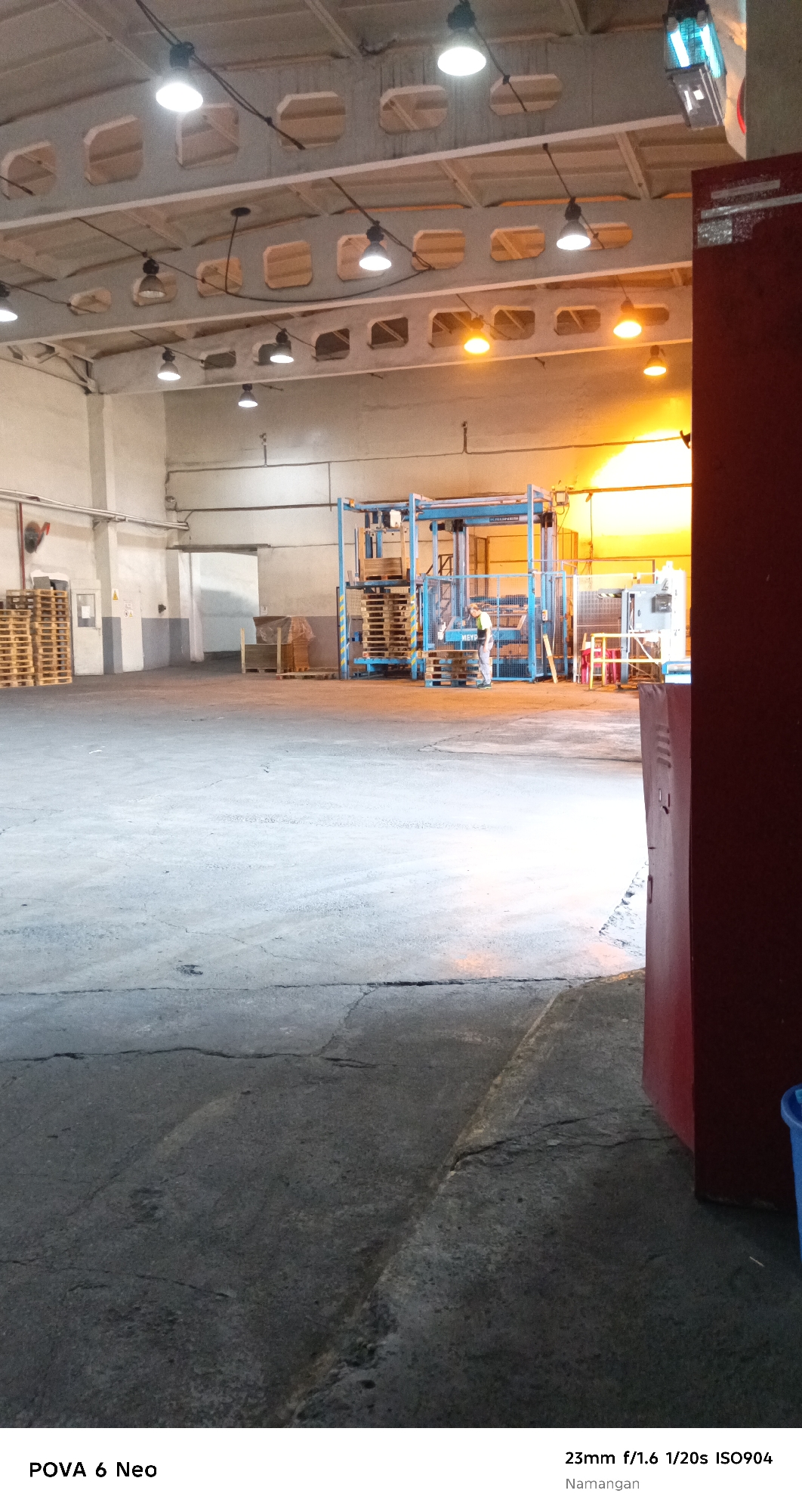 The image depicts the interior of a warehouse in Namangan City, Uzbekistan. The scene is dominated by concrete flooring and a high ceiling with industrial lighting fixtures. Pallets of goods are stacked in various areas of the warehouse, suggesting storage and distribution activities. In the background, a large piece of machinery, possibly for packaging or manufacturing, is visible. A person, wearing a light-colored shirt and darker pants, is interacting with the machinery. The overall impression is one of an operational industrial space. The lighting suggests it might be late in the day, with a warm glow emanating from the background.