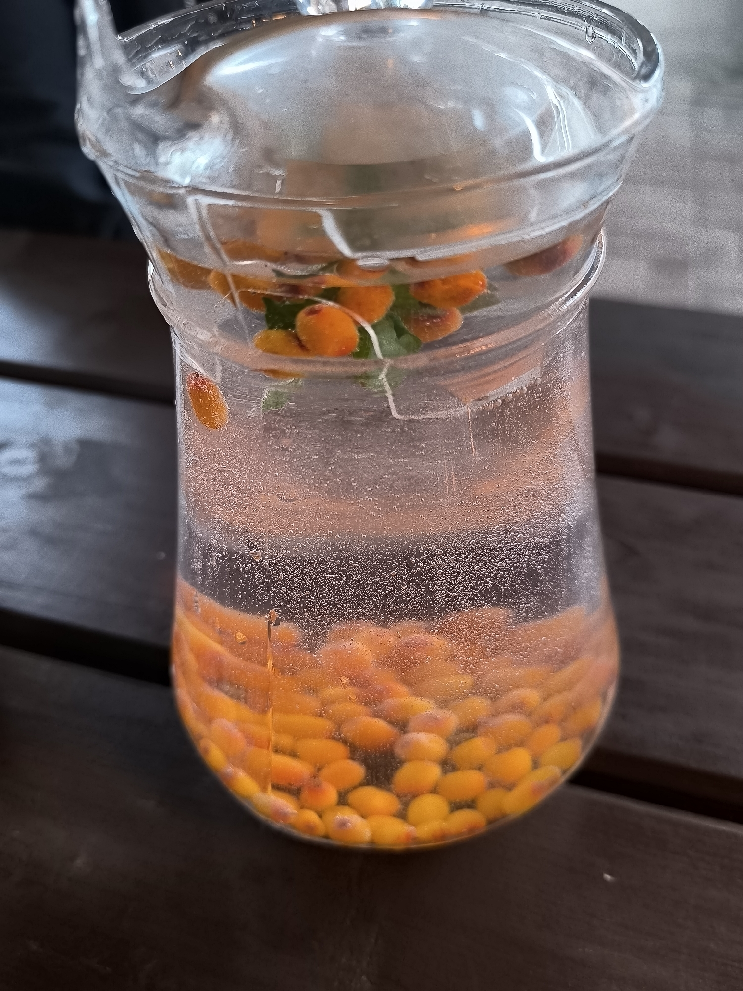 A glass pitcher filled with clear liquid and orange berries, possibly sea buckthorn, sits on a dark wooden table. Some berries are suspended in the liquid, while a large concentration rests at the bottom. Bubbles are visible throughout the liquid, and a few sprigs of green herb are also present. The pitcher's handle is visible on the left side. The background shows a blurred outdoor setting, suggesting this photograph was taken at an outdoor cafe or restaurant. The presence of sea buckthorn berries often indicates a connection to local cuisine or beverages, common in regions like Latvia. The lighting is natural, suggesting daytime.
