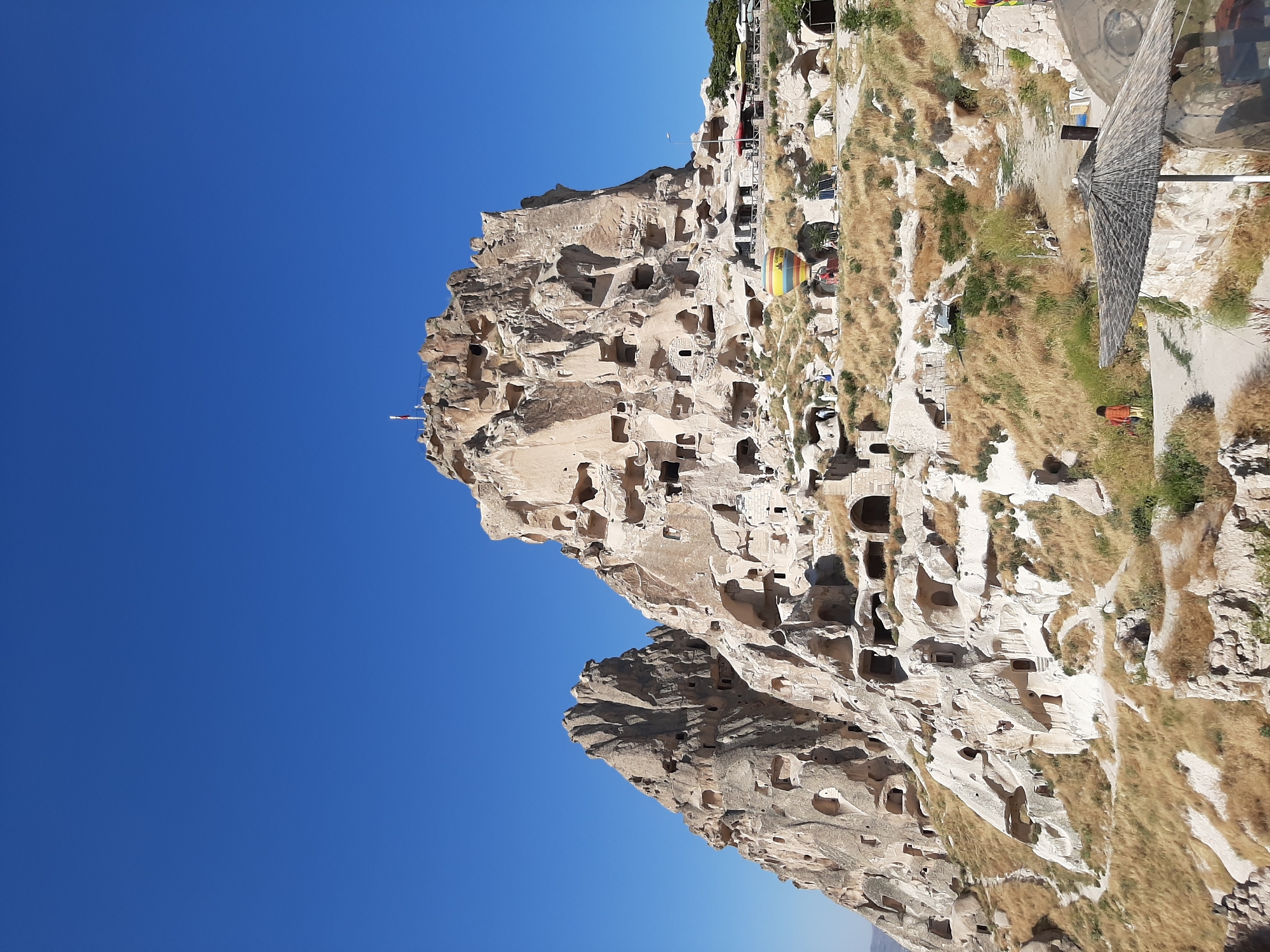 The image depicts Uçhisar Castle, a prominent rock-hewn fortress located in Uçhisar Beldesi, Turkey. The castle is a massive natural rock formation that has been extensively carved with numerous caves and dwellings, giving it a unique, honeycomb-like appearance. The structure rises majestically against a clear, deep blue sky.
In the foreground and mid-ground, the rocky landscape is covered in dry, golden grass and sparse vegetation, indicative of an arid climate. Pathways wind through the terrain, leading towards the castle. Several small structures, some with thatched roofs, are visible at the base of the formation, suggesting tourist facilities or historical remnants.
A hot air balloon, featuring vibrant stripes of yellow, blue, and red, is inflated and visible near the middle of the image, partially obscured by the rocky terrain. A few figures are scattered across the scene, some appearing to be walking along the pathways, including one person in an orange and brown outfit walking towards the left. Another figure is seen near the hot air balloon. A small flag flies from the highest point of the castle. The overall impression is a historical and natural landmark bathed in bright daylight, with signs of human activity and tourism.