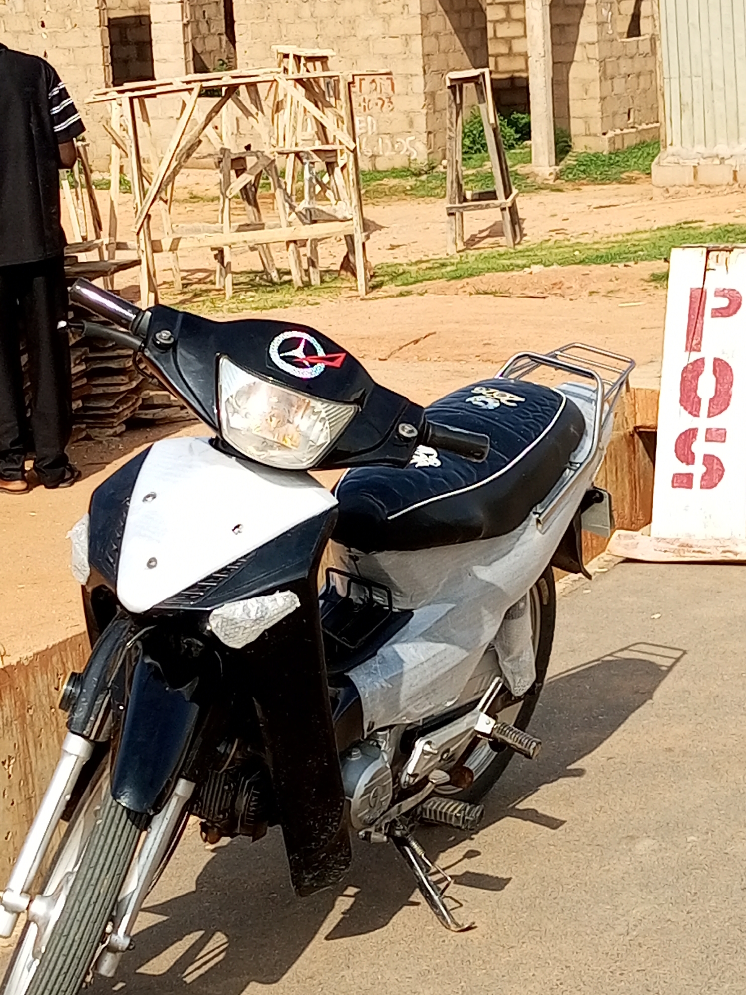 The image shows a parked motorcycle with a black and white front fairing, a black seat with white trim, and a chrome luggage rack. A person wearing a black shirt with white stripes and black pants is partially visible to the left, standing in front of a construction site with wooden scaffolding and unfinished brick buildings. A white sign with red stenciled letters, possibly "POST", is visible on the right side of the image. The ground is a paved surface. The scene appears to be outdoors in a city environment.