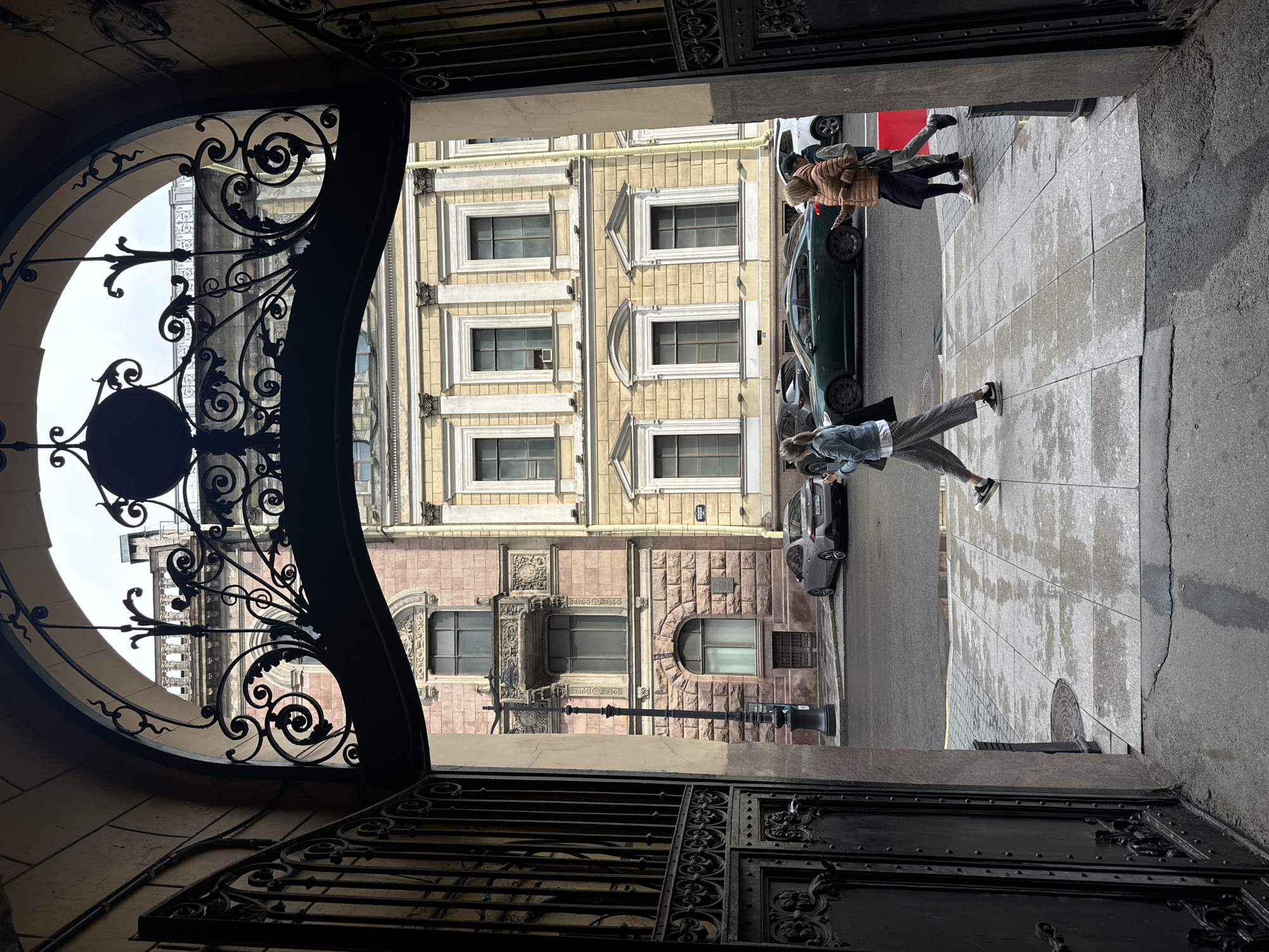 The image captures a view through an ornate, wrought-iron gate into a courtyard or passageway in Saint Petersburg, Russia. The architecture of the buildings visible suggests a historic urban setting. On the left, a building with a pinkish stone facade features classical architectural elements such as columns and balconies. To the right, a building with a yellow facade displays a similar classical style with prominent windows.
In the foreground, the dark, intricate metalwork of the gate dominates the frame, with decorative scrolls and a circular motif. Beyond the gate, the pavement is wet, indicating recent rain. Several people are walking on the sidewalk. On the left, a person wearing a denim jacket and patterned pants walks away from the camera, holding a phone. Further to the right, two individuals are walking towards the camera; one is wearing a brown jacket and the other a grey jacket. A dark green car is parked on the street, with a silver car visible behind it. The sky appears overcast.