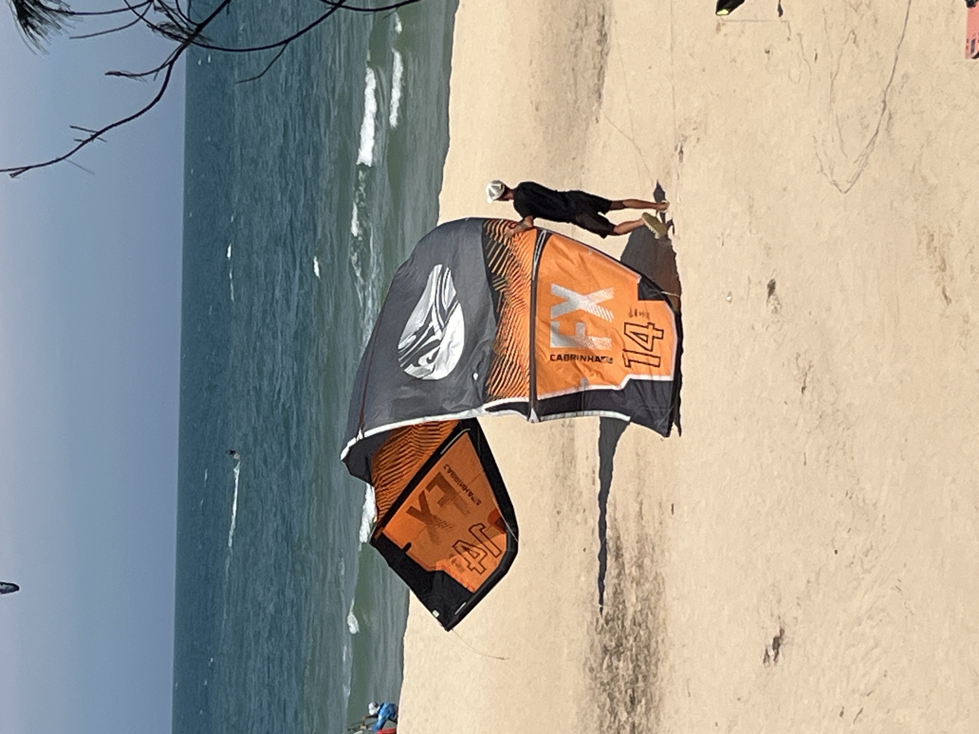 A person in a black t-shirt and shorts, wearing a white cap, stands on a sandy beach in Phu Thuy Ward, Vietnam, holding a large, orange and black kite. The kite, labeled "FX 14", is partially deflated and resting on the sand. The ocean is visible in the background with small waves and a kitesurfer in the distance. A thin tree branch with leaves extends into the upper right corner of the frame. The sky is overcast and the sand appears light brown.