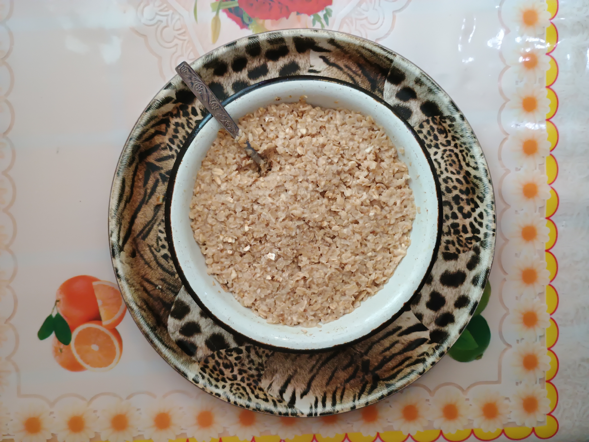A top-down, eye-level, medium shot captures a bowl of oatmeal on a decorative tray. The oatmeal is a light brown color and appears to be cooked with milk. A metal spoon with an engraved pattern rests in the bowl, angled towards the right. The bowl itself is white with a thin black rim. It sits on a round tray decorated with a black and brown leopard print pattern. The tray is on a light pink table with a repeating floral pattern along the edges. In the upper left corner of the image, a small cluster of oranges, one whole and two sliced, are depicted on the tablecloth. The scene is indoors, and there are no people visible. The overall impression is one of a simple meal ready to be eaten.