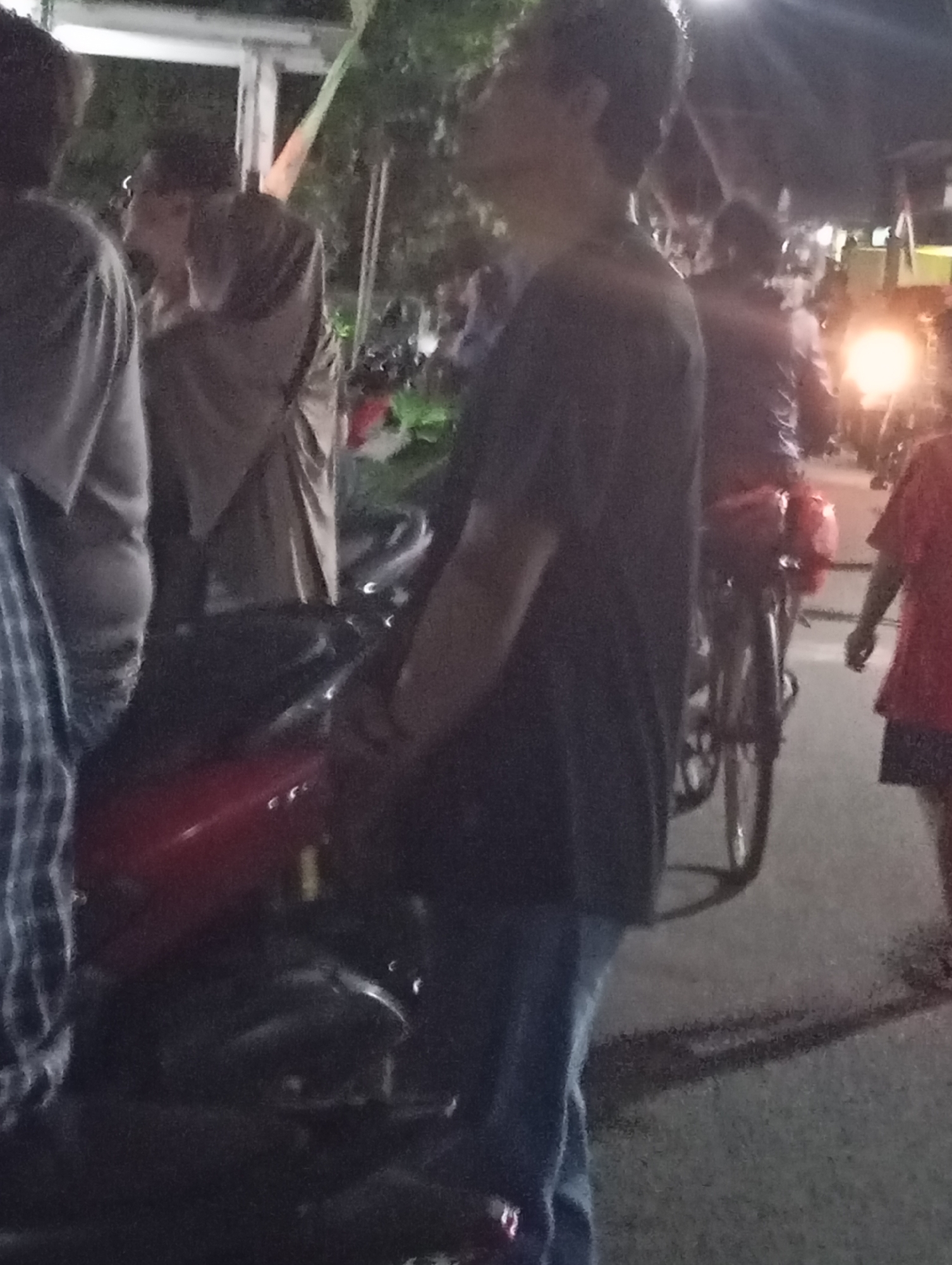 The image captures a nighttime scene on a street in Pacitan, Indonesia, likely during an event or gathering. In the foreground, a person wearing a dark t-shirt and blue jeans stands facing away from the camera, looking upwards. Next to them is a red motorcycle. In the background, other individuals are visible, some with their faces obscured. One person appears to be wearing a light-colored robe or traditional garment. A bicycle with a red carrier is also present. The lighting suggests it is after dark, with artificial lights and possibly streetlights illuminating the area. The environment appears to be a paved street with some foliage in the background.