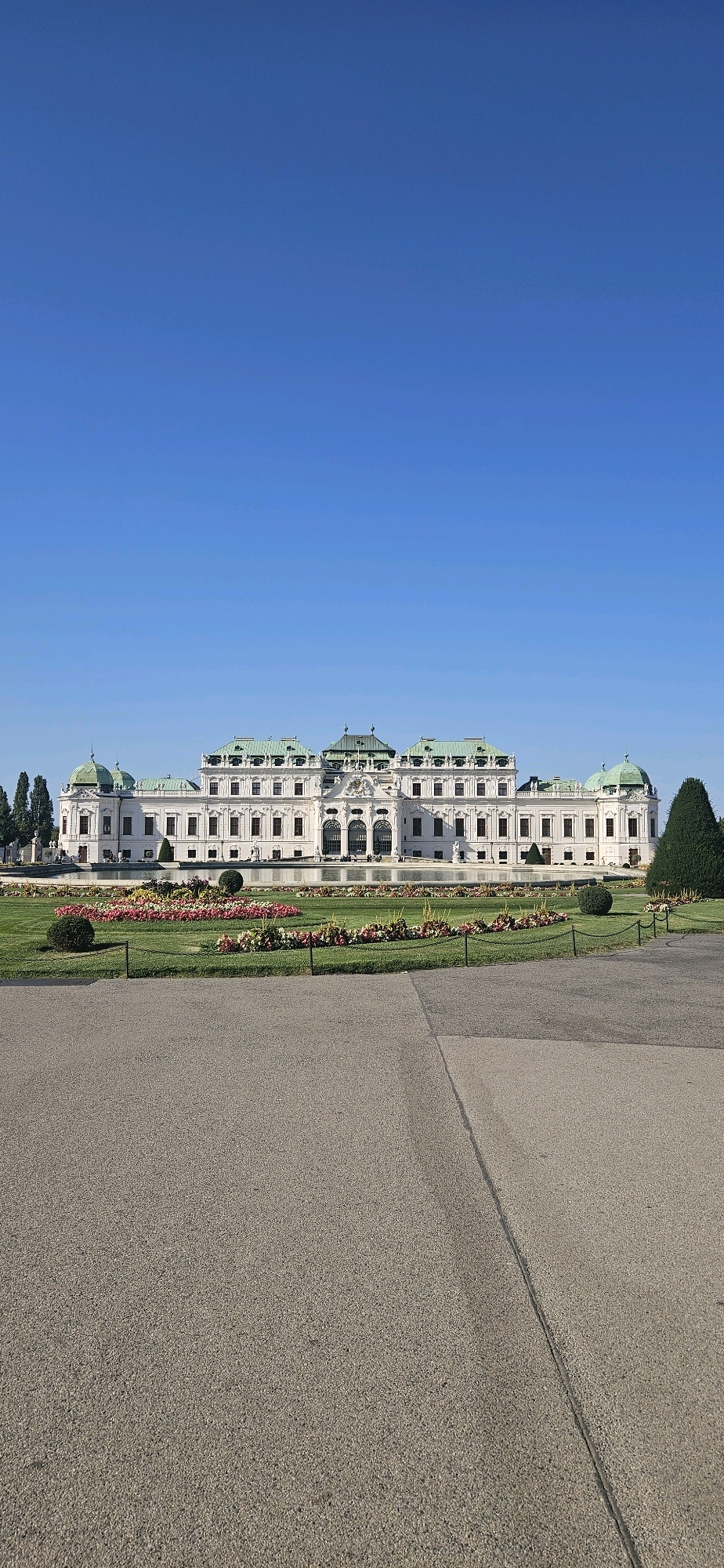 The image shows the Belvedere Palace in Vienna, Austria, on a clear, sunny day. The palace is a grand Baroque complex with a white facade and green domed roofs. It is set against a backdrop of a clear blue sky. In front of the palace, there is a meticulously manicured garden with rows of colorful flowers and neatly trimmed hedges. A pathway leads through the garden towards the palace. The foreground of the image features a paved area, possibly a walkway or parking lot, with a visible seam running down the middle. The overall scene conveys a sense of historical grandeur and natural beauty.
