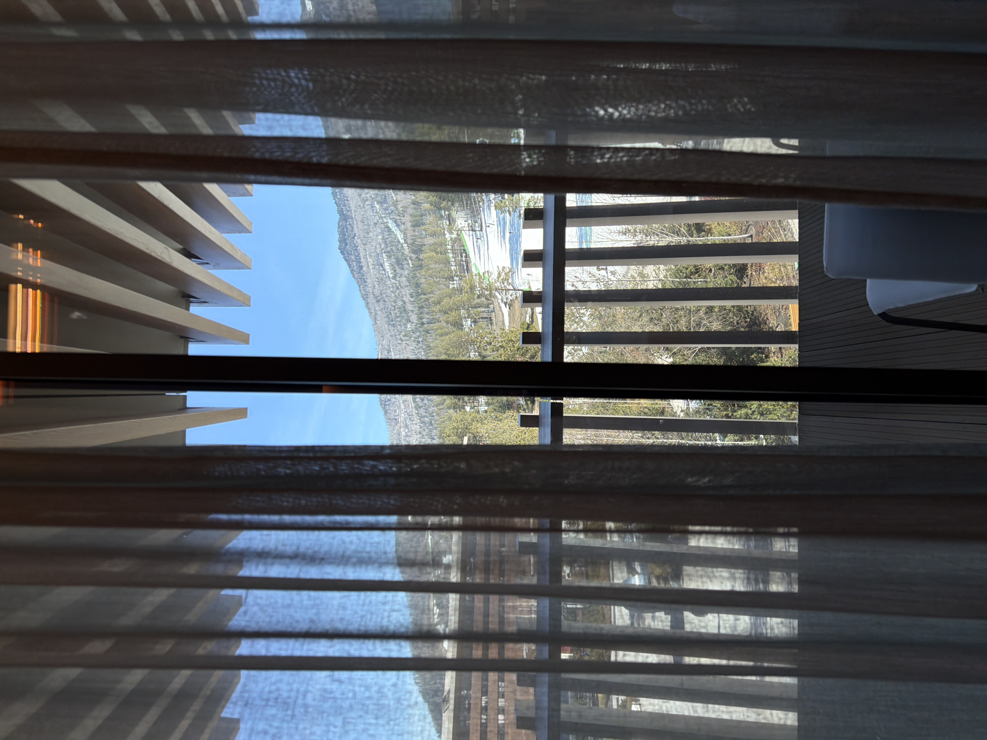 The image shows a view from indoors through a window with semi-transparent curtains and wooden slats. The view is partially obscured by the curtains and slats. Outside, there is a scene of mountains, trees, and possibly a body of water. There is also a balcony or deck visible with a railing and a white chair. It is not possible to determine the location as Озёрное, Russia, without further information.