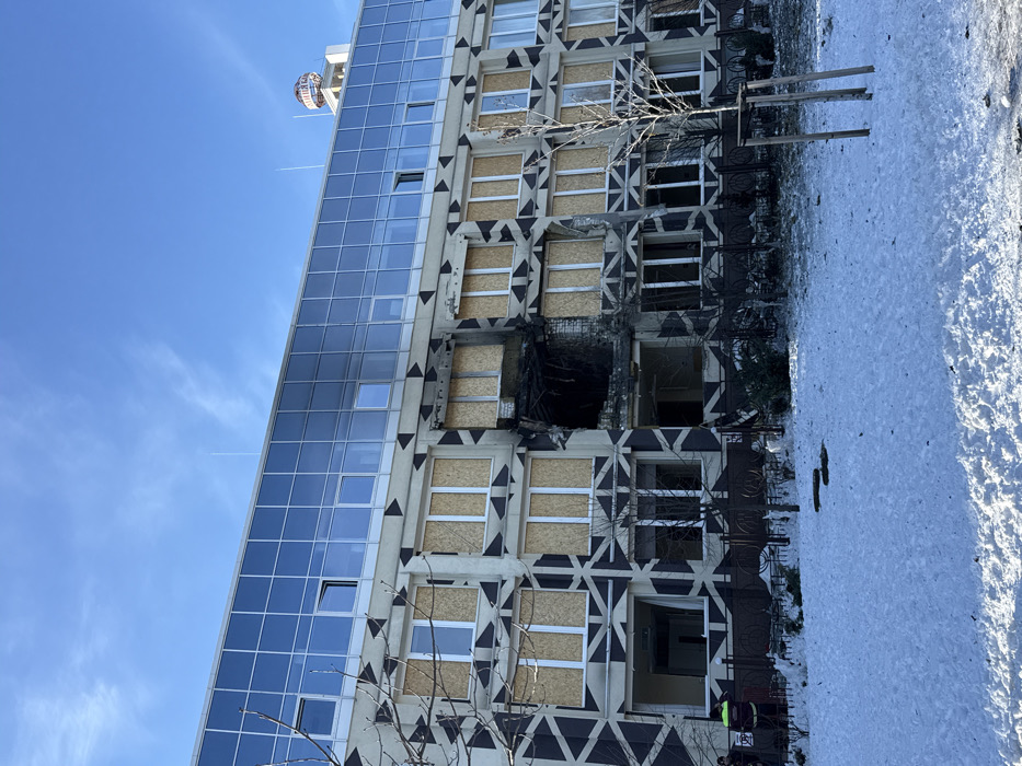 The image displays a multi-story building, identified as the administration building of the Kyiv TV Tower on Dorogozhytska Street, Kyiv, Ukraine, under a clear blue winter sky. A large, irregular breach approximately two stories high is present on the building's facade, centered on the second and third floors from the bottom, exhibiting significant charring and black residue around its perimeter. The lower three visible floors feature a grey facade with a repeating pattern of dark geometric triangles, where most windows are boarded up with light-colored plywood. The upper three floors consist of a modern glass curtain wall, with some panels appearing damaged or open, and smoke discoloration visible on the top-left section. The ground in the foreground is covered in snow, with bare deciduous trees and a black metal fence running along the building's base. Two individuals, one in a high-visibility vest, are partially visible on the far left near the building's foundation. A taller structure with a circular sign displaying the word "КИЕВ" (Kyiv) in Cyrillic is visible behind the damaged building.