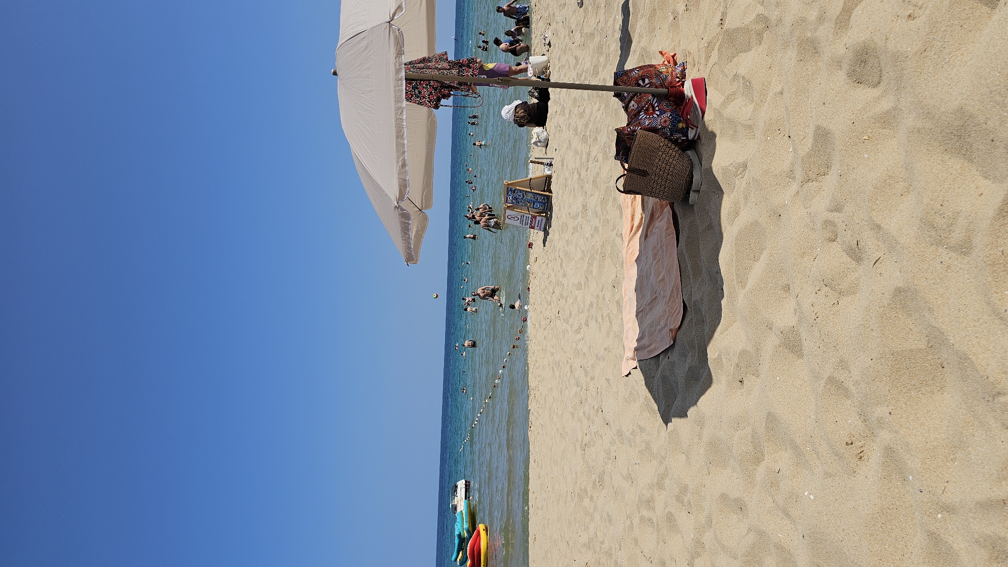 A bright, sunny day at the beach in Varna, Bulgaria is depicted. The scene is divided into sand, sea, and sky. The foreground is dominated by light brown sand, with subtle ripples and footprints. A pale orange beach towel is laid out on the sand, with two bags and a pair of red sandals resting near its edge. A large, light-colored beach umbrella casts a shadow over a portion of the sand and the items placed beneath it.
In the middle ground, the sea stretches out, with a clear blue sky above. People are visible in the water, some swimming, others standing. A rope barrier is strung across the water, demarcating an area. Further out, on the sea, several inflatable watercraft are present, including a yellow and red banana boat and what appears to be a similar watercraft. Two small white motorboats are also visible.
The background is a vast expanse of clear, bright blue sky. The overall impression is one of a typical summer day at a popular beach.