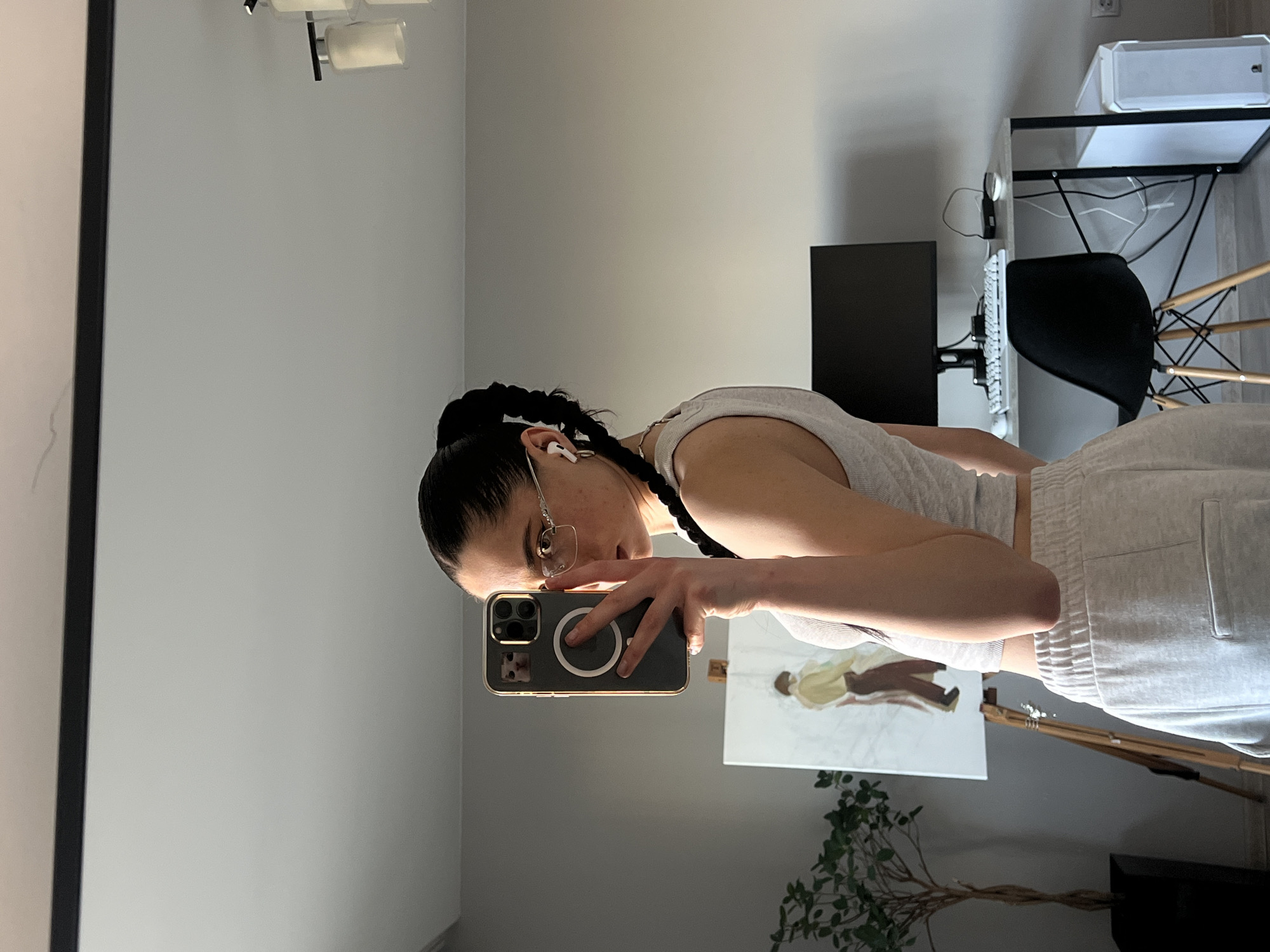 A young woman is taking a selfie in a mirror. She is wearing a light grey crop top and light grey sweatpants. Her hair is pulled back in a ponytail with two braids. She is wearing glasses and white wireless earbuds. In the reflection of the mirror, there is a painting on an easel of two figures, a plant in a black pot, a desk with a computer monitor, keyboard, mouse, and a white computer tower. The room appears to be a home office or studio. There is a light fixture visible on the ceiling. The image is taken in Ternopil, Ukraine.