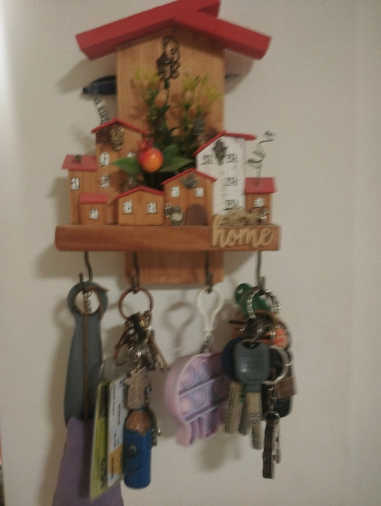 The image shows a wooden key holder shaped like a small village with red roofs, mounted on a white wall. The key holder has several hooks on its bottom edge. Hanging from the hooks are several sets of keys. One set has a blue and brown cylindrical charm with a smiling face. Another set includes a light pink pop-it toy. A purple fabric item is also attached to one of the hooks. The key holder itself is adorned with miniature houses, a faux apple, and greenery. The text "home" is carved into a wooden plank on the key holder. This scene is likely a domestic setting in the city of Ashdod, Israel, possibly a household entryway.