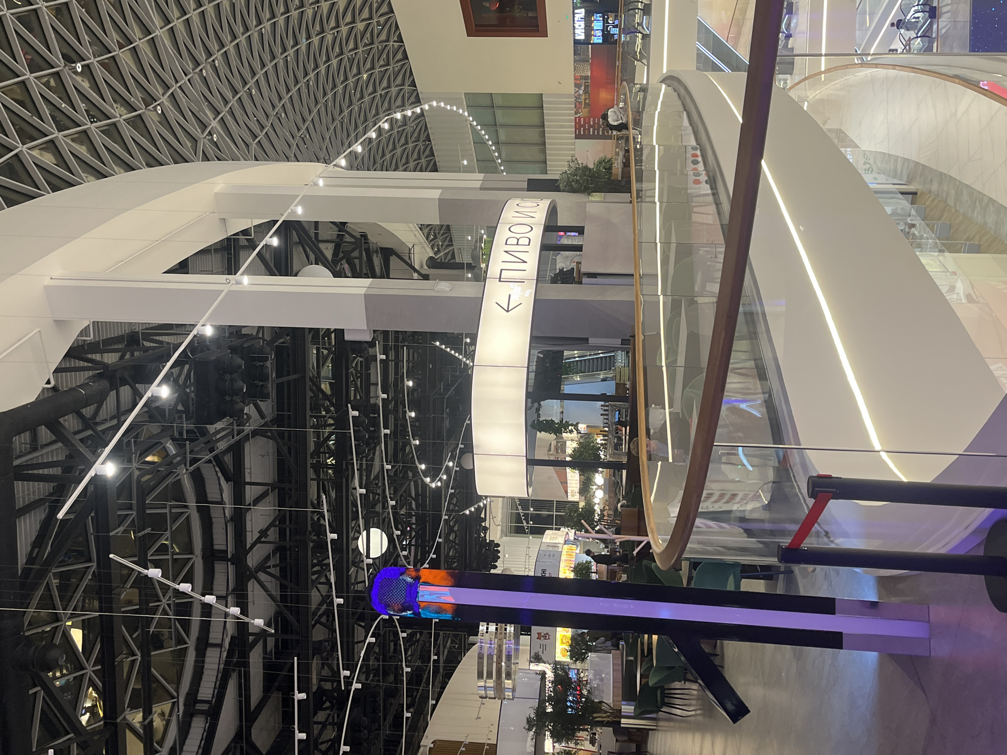 The image shows the interior of a large shopping mall in Saint Petersburg, Russia, identifiable by the Cyrillic sign reading "ПИВО И СИ" (Pivo i Si), which likely indicates a location selling beer and cider. The mall features a high, arched ceiling with a complex metal framework. Several levels are visible, connected by escalators with glass railings. There's a directional sign pointing towards the beer and cider location. Below, a vertical display, possibly a digital screen, shows colorful patterns and advertising content. Tables and chairs are visible in a seating area, suggesting food vendors are nearby. The mall appears modern and well-lit.