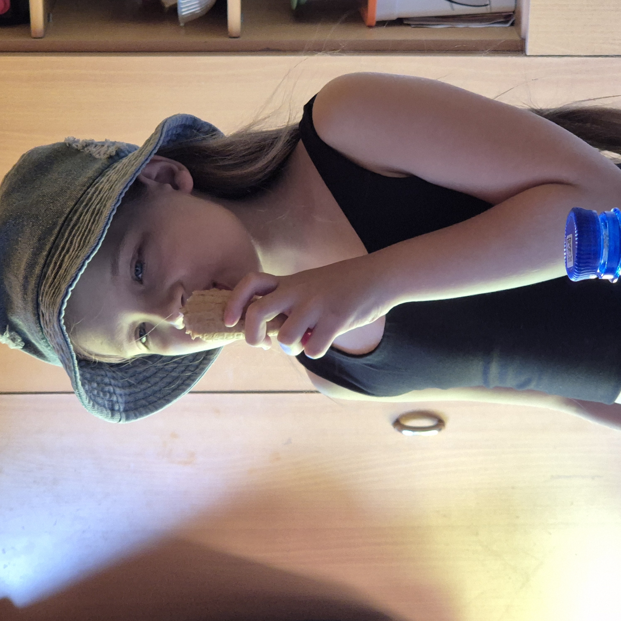 A young person, likely a child or pre-teen, with long, dark hair, is shown in a portrait-oriented image that appears rotated 90 degrees clockwise. They are wearing a black tank top and a distressed denim bucket hat, and are engaged in eating a light-colored, rectangular waffle. The person's left hand holds the waffle to their mouth, while their light-colored eyes (blue or green) look towards the camera with a neutral expression.
The setting appears to be indoors, possibly a home in Новостройка, Russia, with the subject lying or leaning on a light-colored wooden surface. In the upper right corner of the original, unrotated image (which corresponds to the right side of the current image), a blue plastic bottle with a cap bearing faint, unreadable text is visible. A golden ring rests on the wooden surface below the person's arm. The background includes glimpses of shelves or wooden furniture. The lighting suggests an indoor source, casting a gentle glow on the subject and the waffle.