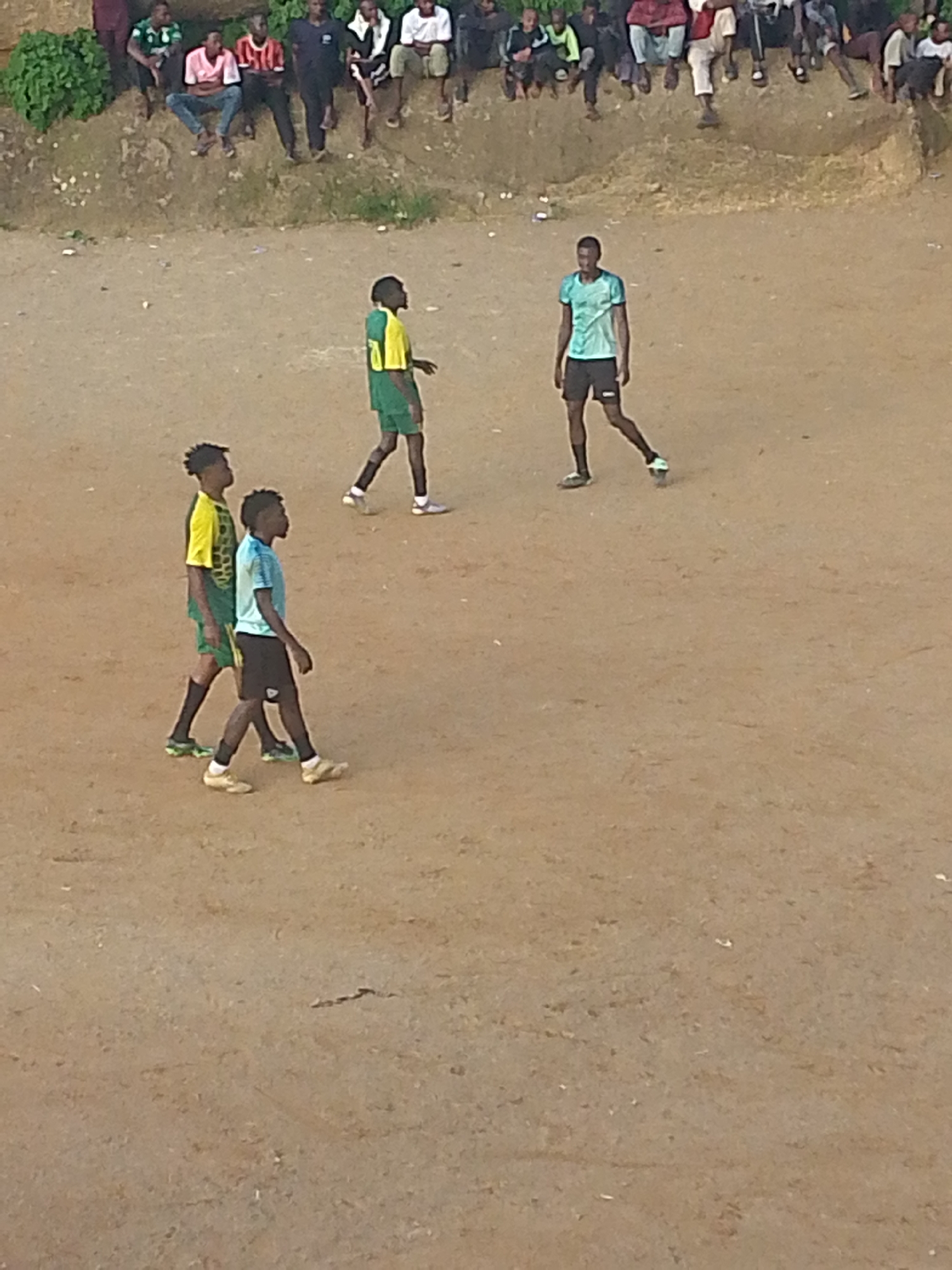 The image depicts an outdoor scene with several young men engaged in a game of football on a dirt field. The men are wearing athletic attire, including jerseys and shorts, suggesting a casual match. Some spectators are visible in the background, seated on a raised embankment. The environment appears to be a public space, possibly a local park or a community field within the city of Jos, Nigeria. The ground is dry and dusty, typical of the terrain in some parts of Jos. The overall atmosphere seems informal and recreational, with the focus on the game and the camaraderie of the participants.