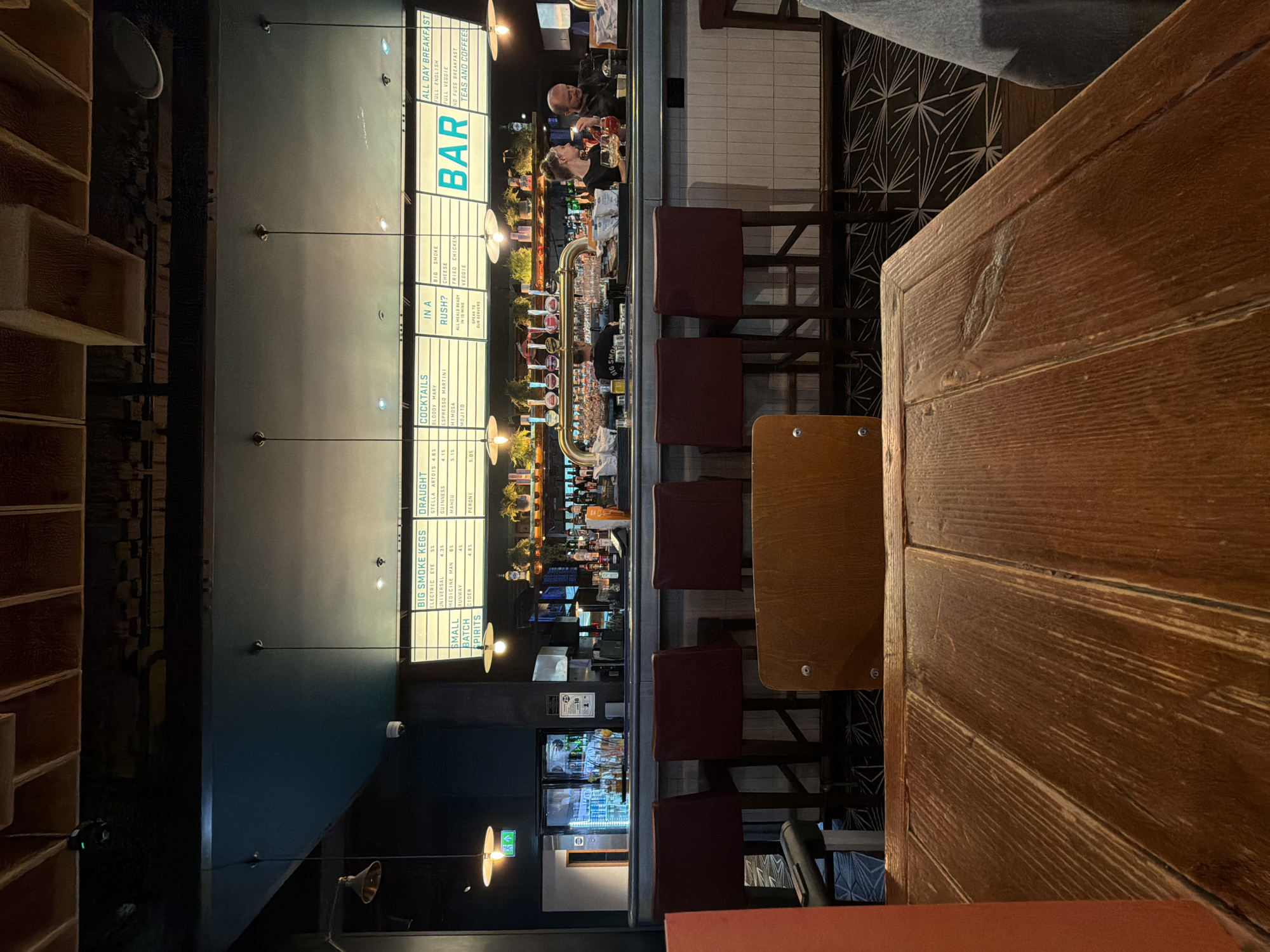This media file captures the vibrant interior of a modern bar or pub, likely located in Crawley, United Kingdom. The scene is viewed from a seated perspective at a wooden table, looking across a long bar counter towards the service area.
The central subject is the well-stocked bar, illuminated by several pendant lights. Behind the counter, two individuals, presumably staff, are engaged in preparing drinks or serving customers. One person with a beard is visible further down the bar, while another with dark hair is leaning over the counter near the beer taps. The bar features an elaborate multi-tap beer system, indicating a variety of draft options. A line of dark red upholstered bar stools is positioned in front of the counter for patrons.
A prominent, illuminated menu board dominates the wall behind the bar, displaying various offerings. To the left, wooden shelving units add to the bar's decor. In the foreground, the wooden table from which the photo was taken shows a textured surface. The flooring beneath the bar stools features a distinctive geometric pattern. The overall lighting suggests it is evening or night, contributing to a lively and social atmosphere.
Visible text on the menu board includes:
- "BAR"
- **ALL DAY BREAKFAST:** "FULL ENGLISH", "FULL VEGGIE", "NO FUSS BREAKFAST", "TEAS AND COFFEES"
- **COCKTAILS:** "BLOODY MARY", "ESPRESSO MARTINI", "MIMOSA", "MOJITO", "SPEAK TO SERVERS"
- **IN A RUSH?:** "ALL MEALS READY IN 10 MIN"
- **BIG SMOKE:** "CHEESE", "FRIED CHICKEN", "VEGGIE"
- **BIG SMOKE KEGS:** "ELECTRIC EYE 5%", "UNIVERSAL 4.3%", "MEDICINE MAN 6X", "RUNWAY 4%", "CIDER 4.8%"
- **DRAUGHT:** "STELLA ARTOIS 4.6%", "GUINNESS 4.1%", "MAHOU 5.1%", "PERONI 5.0%"
- Partially visible text for "SMALL BATCH SPIRITS".