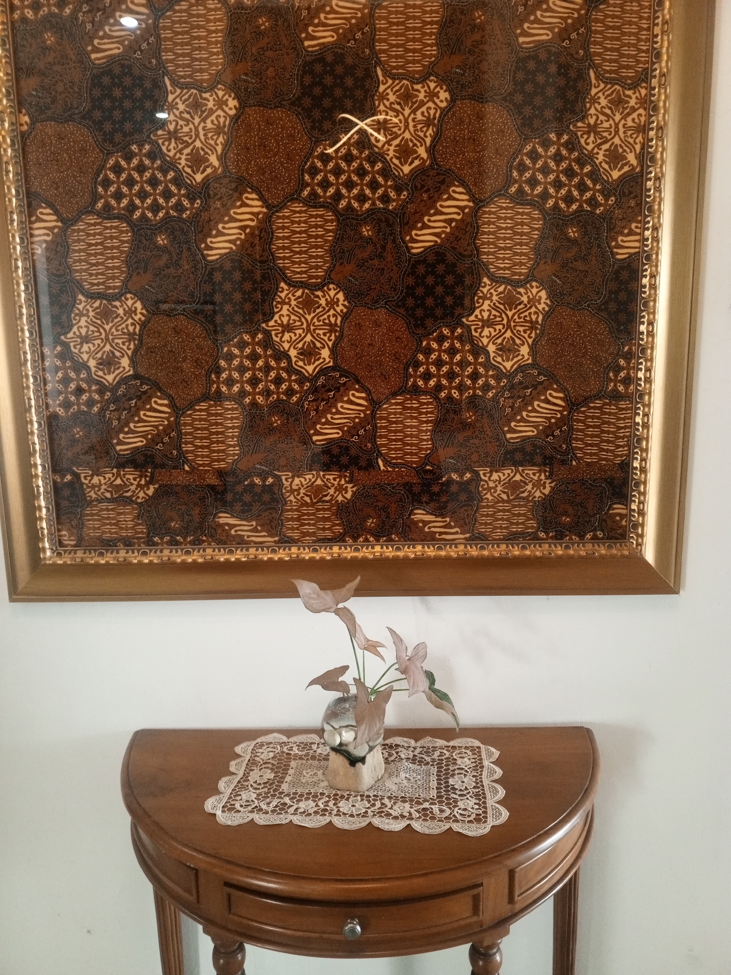 A framed batik artwork hangs on a white wall above a half-moon shaped wooden table. The batik features a repeating pattern of organic shapes in shades of brown and cream, with a golden frame. On the table, a delicate lace doily is placed, and on top of the doily sits a small vase with a plant and a few pebbles. The table has a single drawer with a small silver knob. The scene appears to be indoors, in a well-lit room, possibly a home or a gallery. There are no people visible in the image. The image is static and does not depict any actions or interactions. The location is identified as Bandung City, Indonesia.