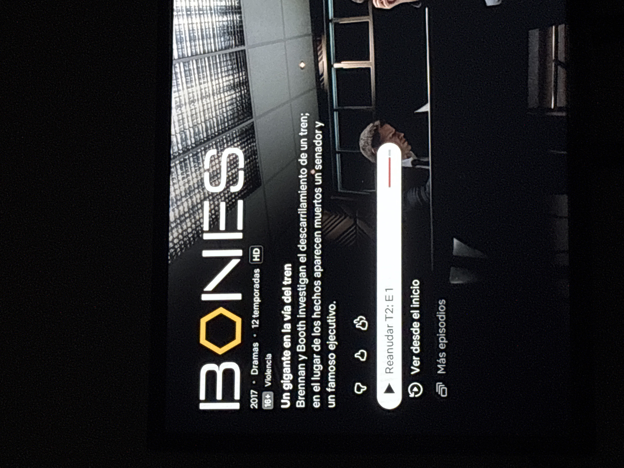 A slightly rotated image of a television screen displays the title card of the television show "Bones". The title "BONES" is prominently displayed with the "O" replaced by a yellow hexagon. Below the title, details about the show are listed: "2017 Dramas 12 temporadas HD", "16+ Violencia". A brief synopsis is provided in Spanish: "Un gigante en la via del tren; Brennan y Booth investigan el descarrilamiento de un tren; en el lugar de los hechos aparecen muertos un senador y un famoso ejecutivo."
Beneath the synopsis, there are playback options: "Reanudar T2: E1" (resume season 2, episode 1) and "Ver desde el inicio" (watch from the beginning). A "Más episodios" (more episodes) option is also visible. In the background, there are blurred images of buildings and a man's face.