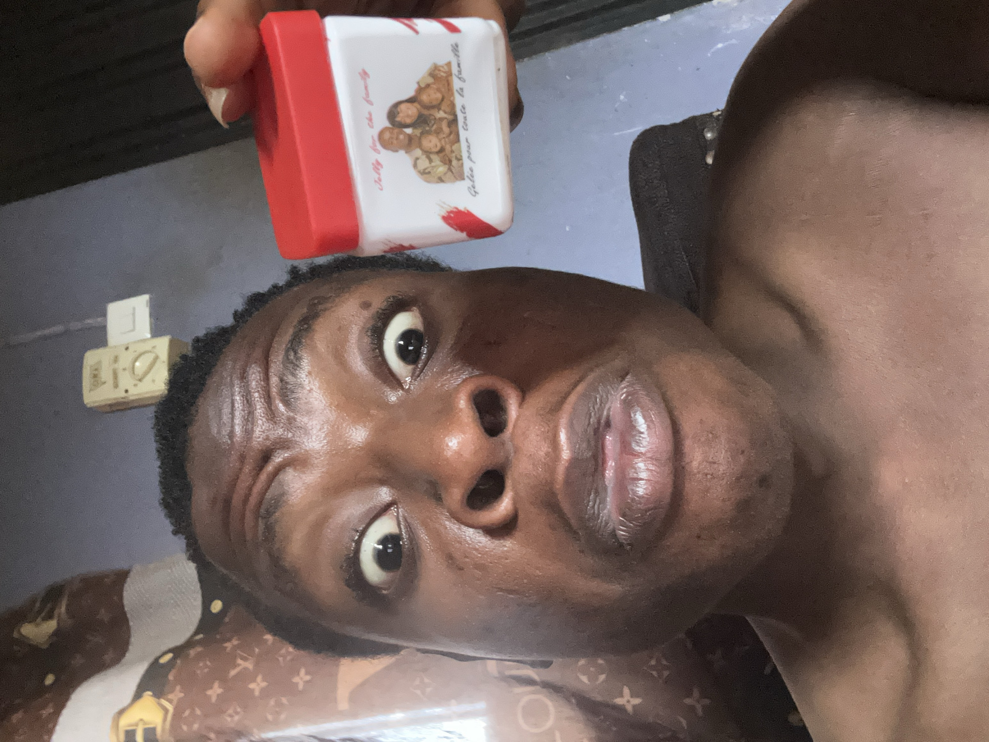 A shirtless person of African descent is holding a red and white container with a picture of a family on it. The container says "Jolly for the family" and "Galée pour toute la famille". The person is looking directly at the camera. The background is blurred, but a bed with a brown and gold pattern can be seen. An electrical outlet is visible on the wall. The location and city are unknown.